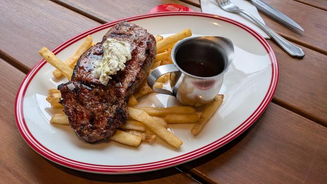 an oval plate with a pile of thin chips. Sitting on top is a charred steak topped with a dollop of tarragon butter. To the side is a little jigger of gravy