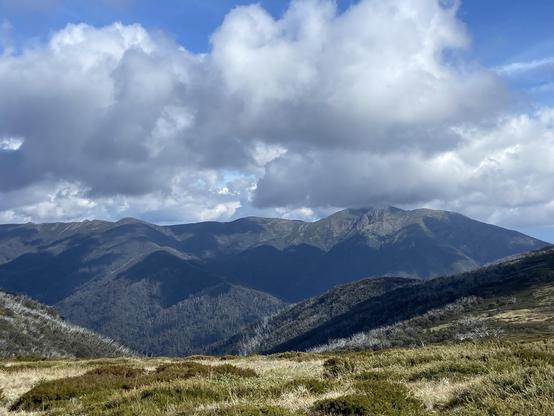 A view of mountains under a partly cloudy sky, with varying shades of green and brown vegetation in the foreground and peaks in the distance.