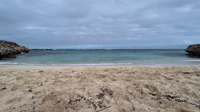 A narrow, sandy bay with rocky sandstone headlands either side. Mostly clear, turquoise water gently breaks on the sand. The sky is mostly overcast and heavier surf breaks over reefs further out.
