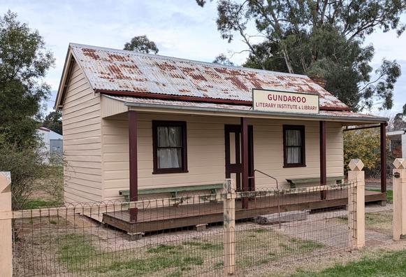 A small 2 window, 1 door slat walled building with a rusting galvanised iron roof. The sign over the verandah reads "Gundaroo literary institute and library".