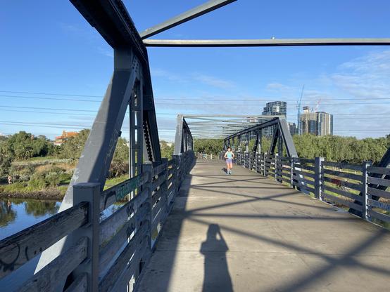 A runner makes her way across a footbridge over a river under blue skies with some tall buildings in the background