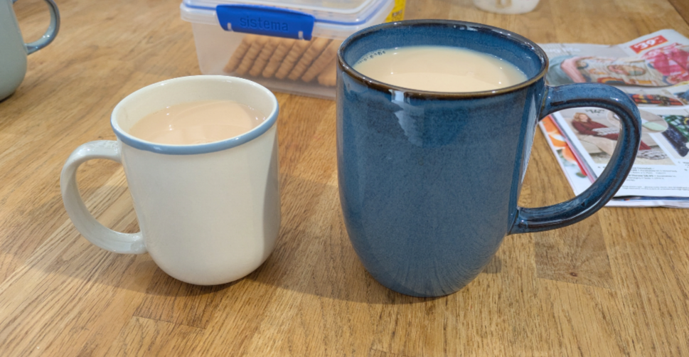 A photograph of two coffee mugs sitting on a wooden table. There is a pale coloured one with a blue rim on the left, and on the right is a blue glazed mug that is at least 50% larger in every dimension. Both are filled with tea.