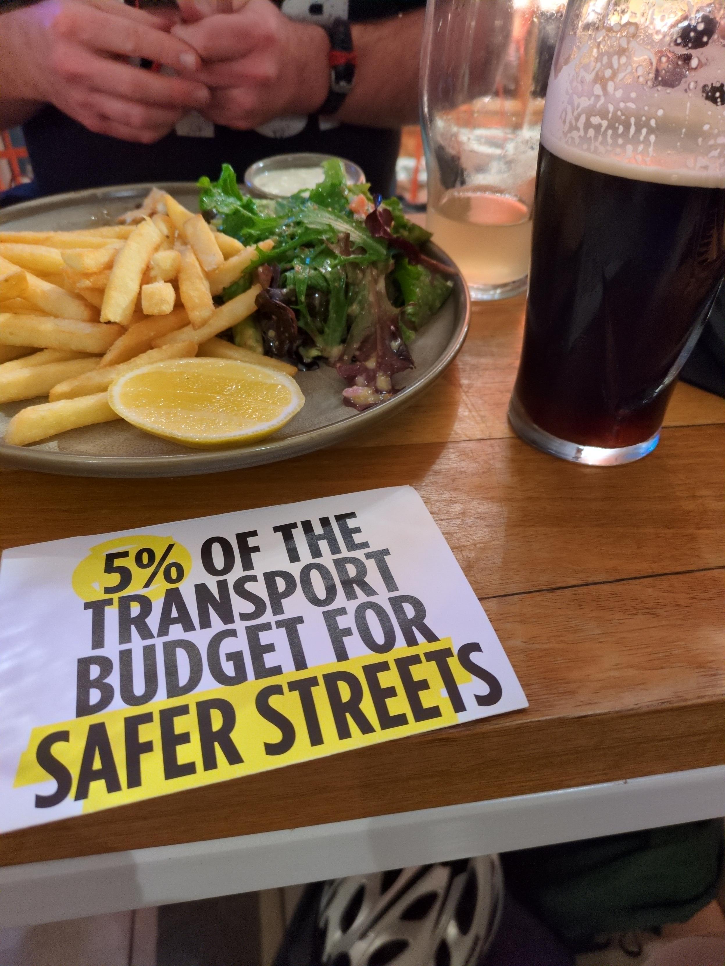 A photo of a leaflet for "5% of transport budget for safer streets" on a table next to a pint of beer and pub chips and salad.
