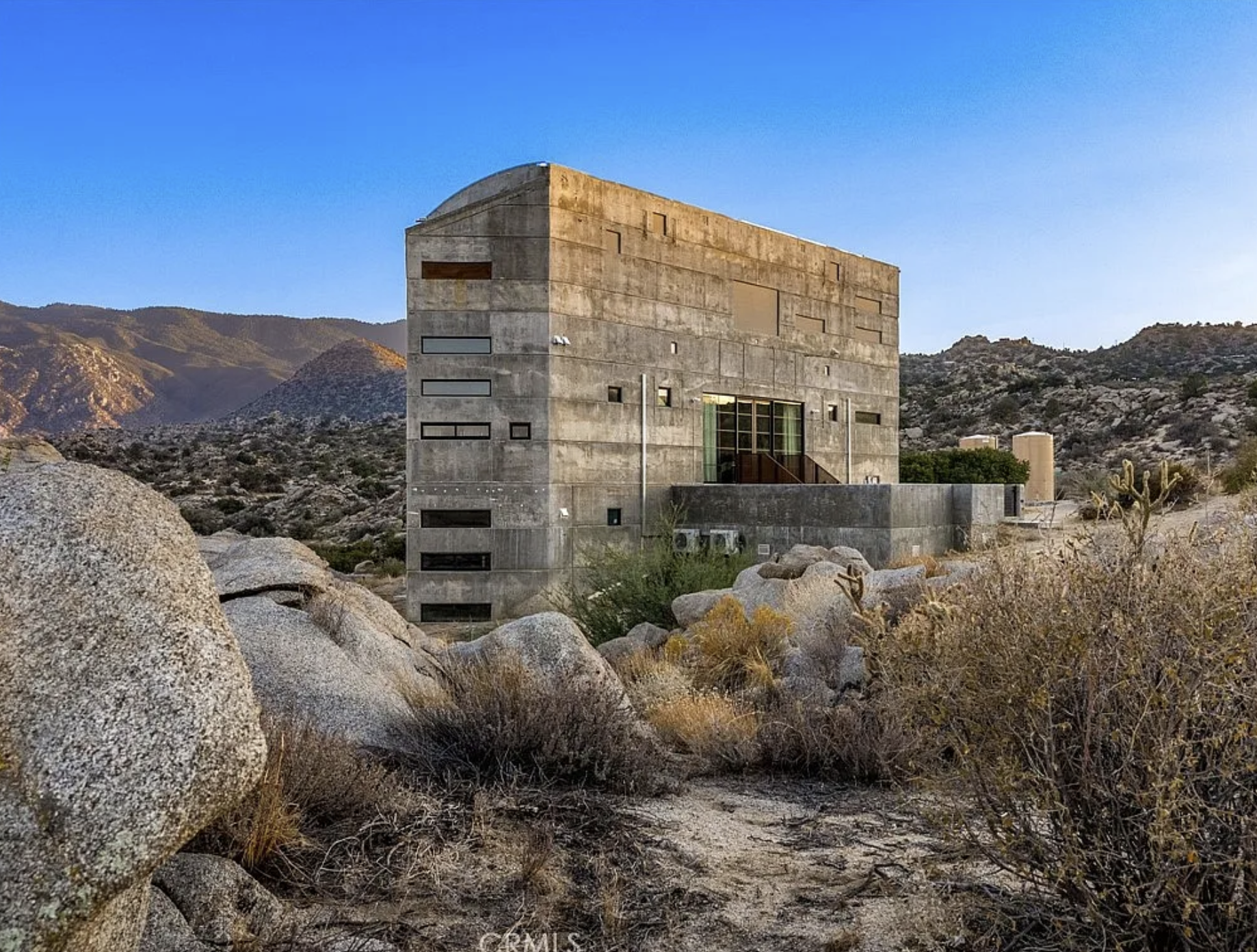 A wide monolith looking house that is several stories high, and clad entirely in concrete. It is standing by itself in a mountainous desert landscape.