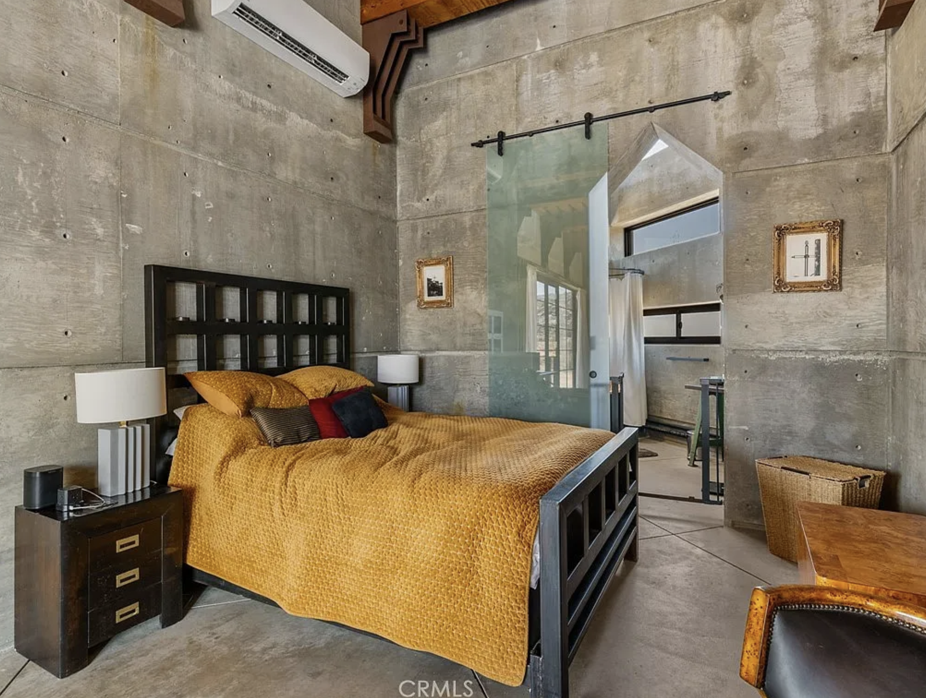 A bedroom inside the house, and all the walls are concrete with a glass sliding door towards the next room.