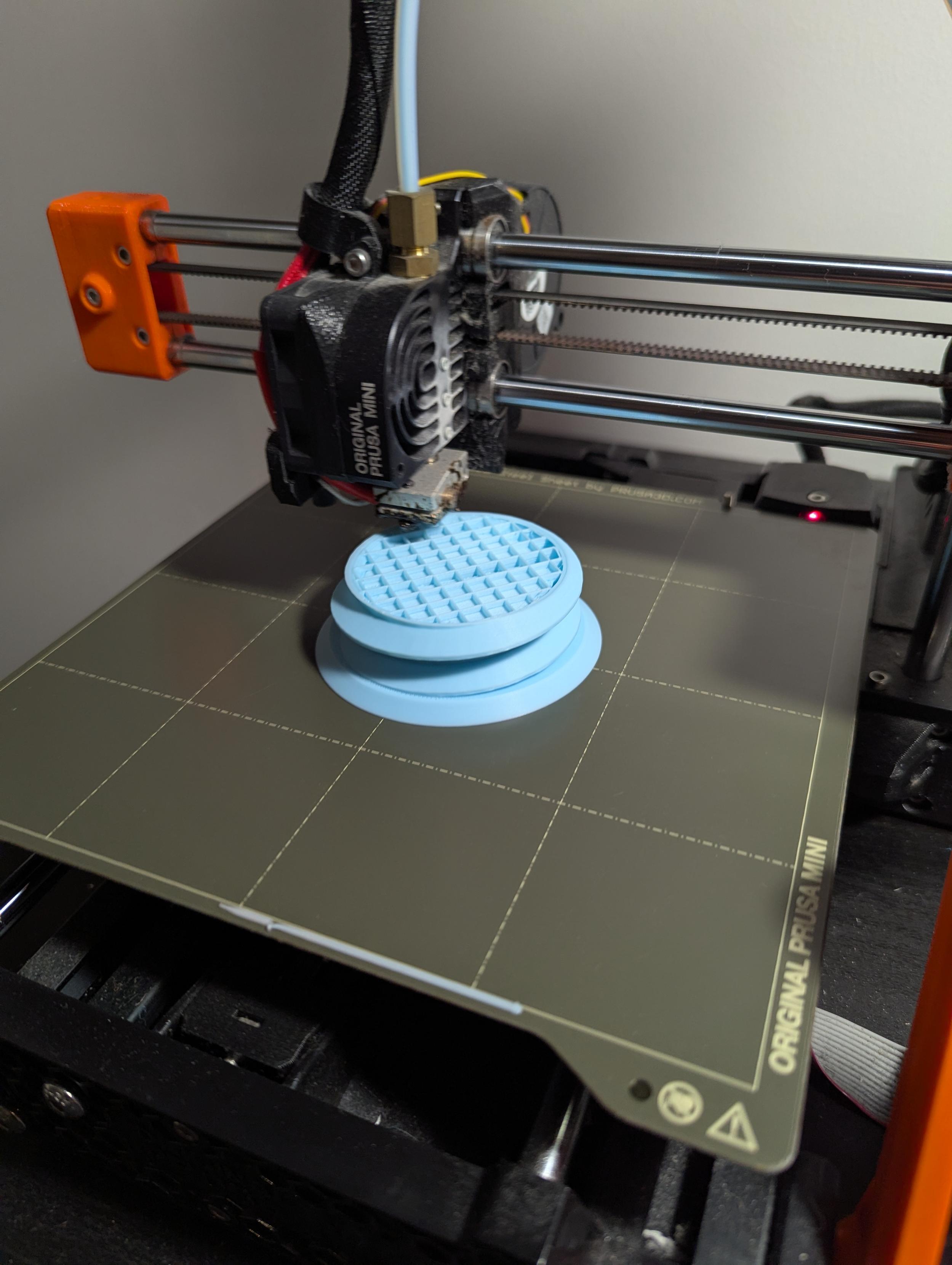 Photo of a Prusa Mini+ 3D printer part way through printing something in pale blue matte finish filament. The thing has a large thread structure on the outside edges.