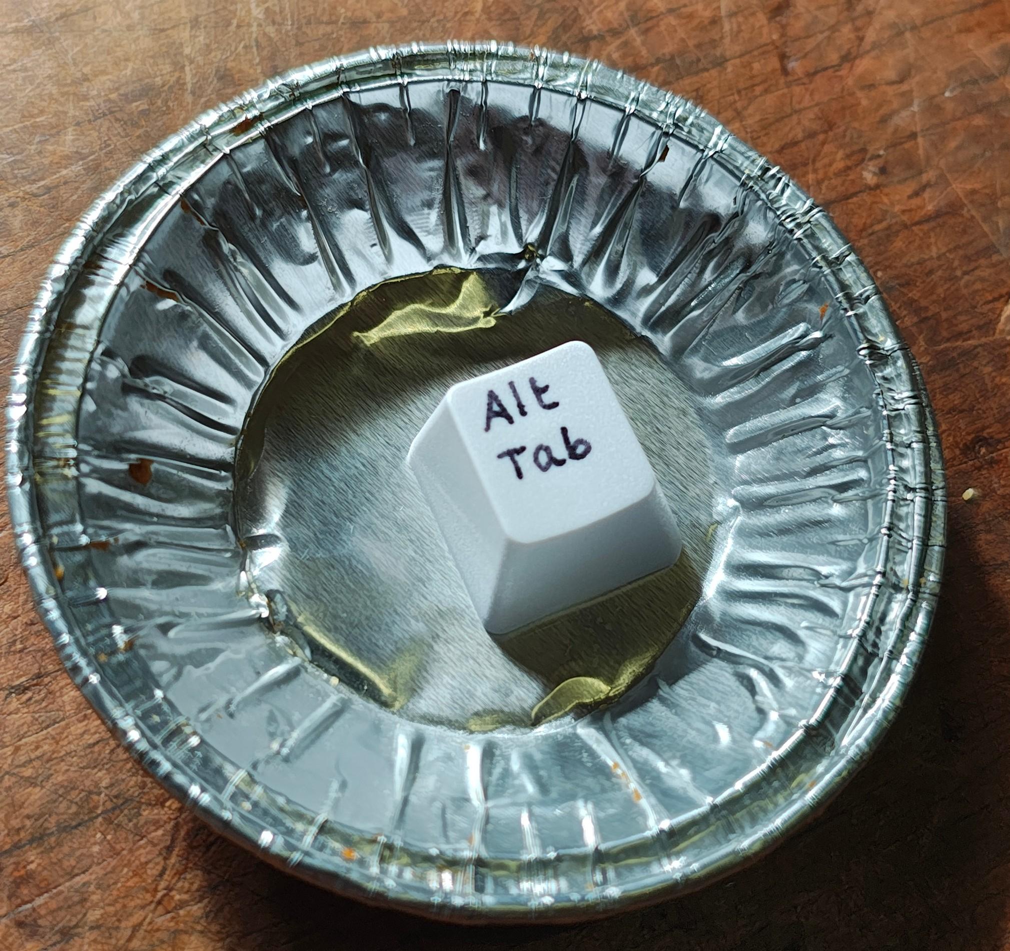 A white computer key cap with the text "Alt Tab" handwritten in blank. It's in a small aluminum tray.