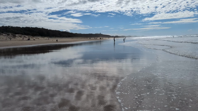 Walking along Coolum Beach while the tide is out. The flat sand is mirror-like as water seeps-out from underground and flows toward the ocean edge while low waves roll the other way over it.