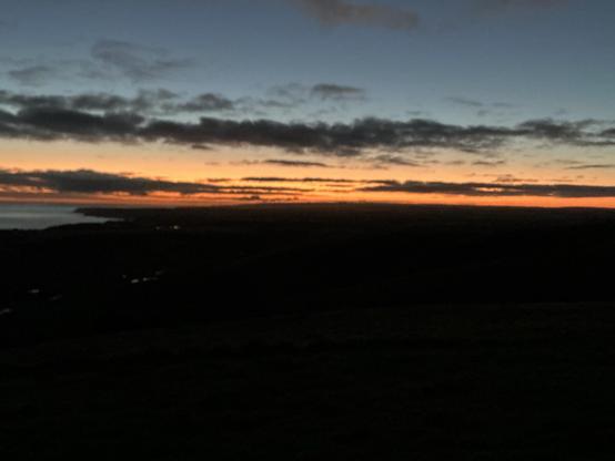 A landscape featuring a colorful sunrise with shades of orange and blue. The horizon line stretches across the image, showcasing a coastal view with clouds scattered in the sky. Silhouettes of rolling hills are visible in the foreground.