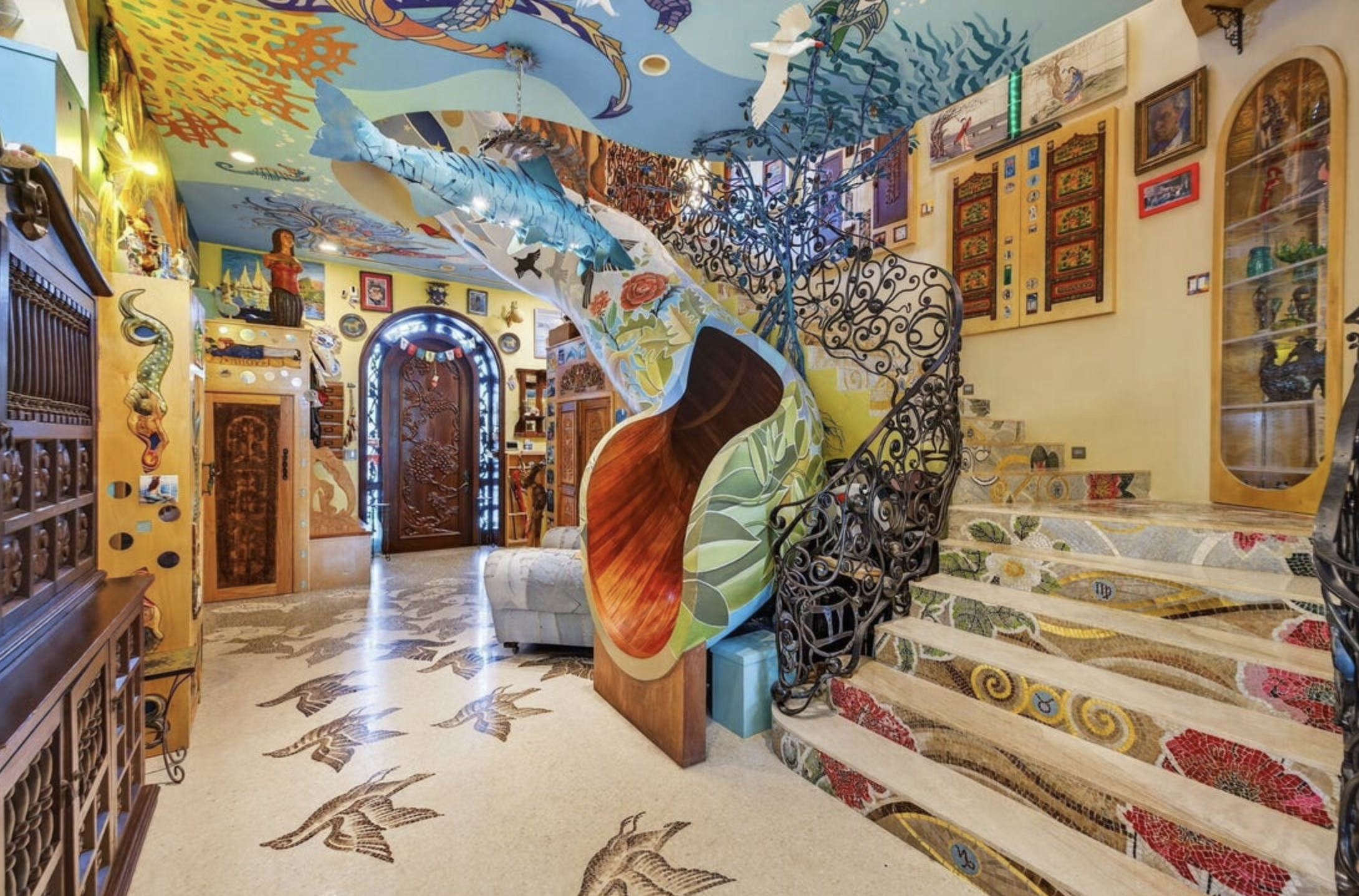 The foyer of a house with a plethora of different patterns across the floor, steps, ceiling, and there is a large brightly coloured slide next to the steps, where one could bypass it and go down from the next floor.