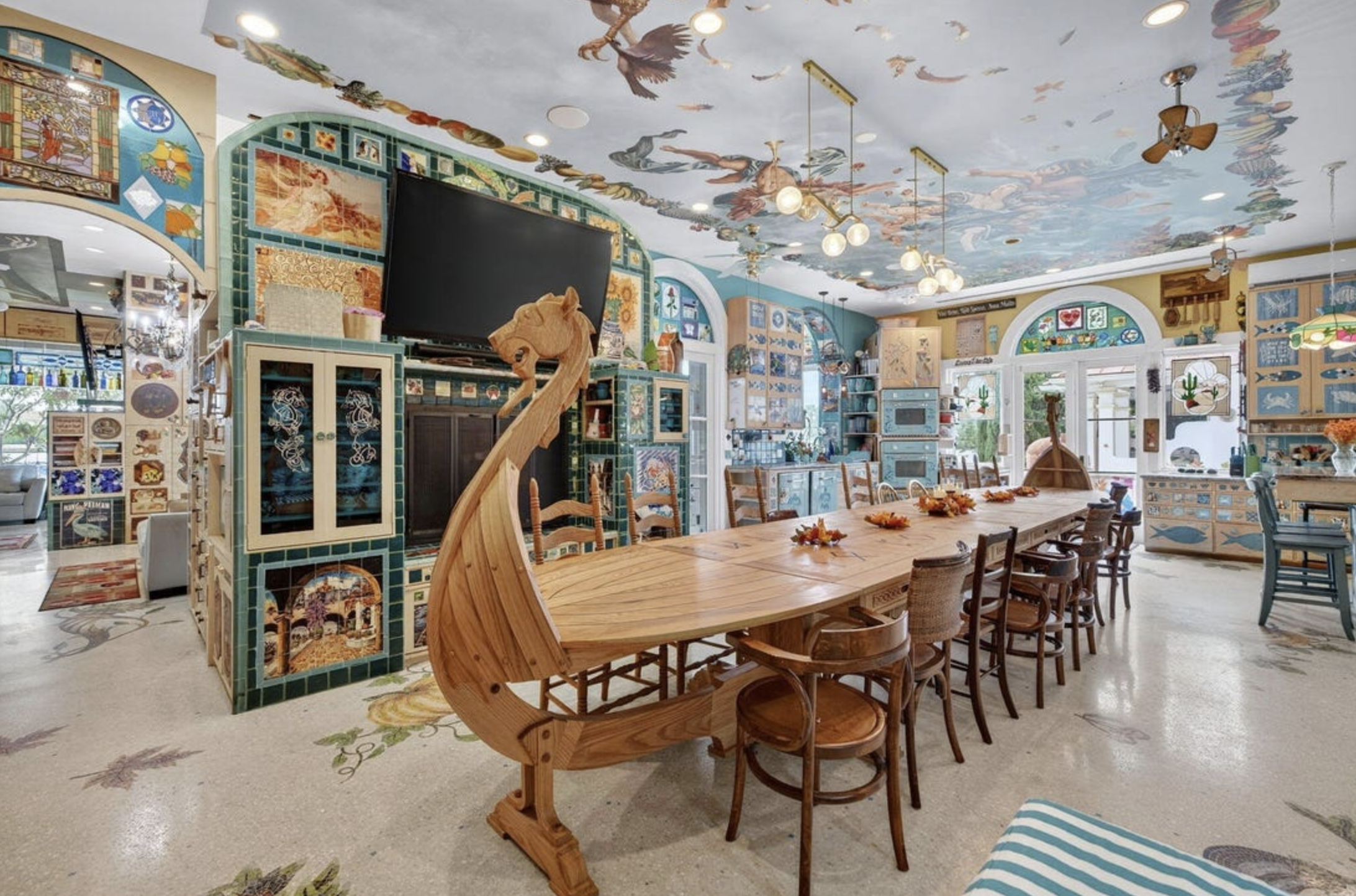 A dining room with a very long wooden table with the head of it shaped like a Viking ship. There are all different patterns on the ceiling, wall and floors.
