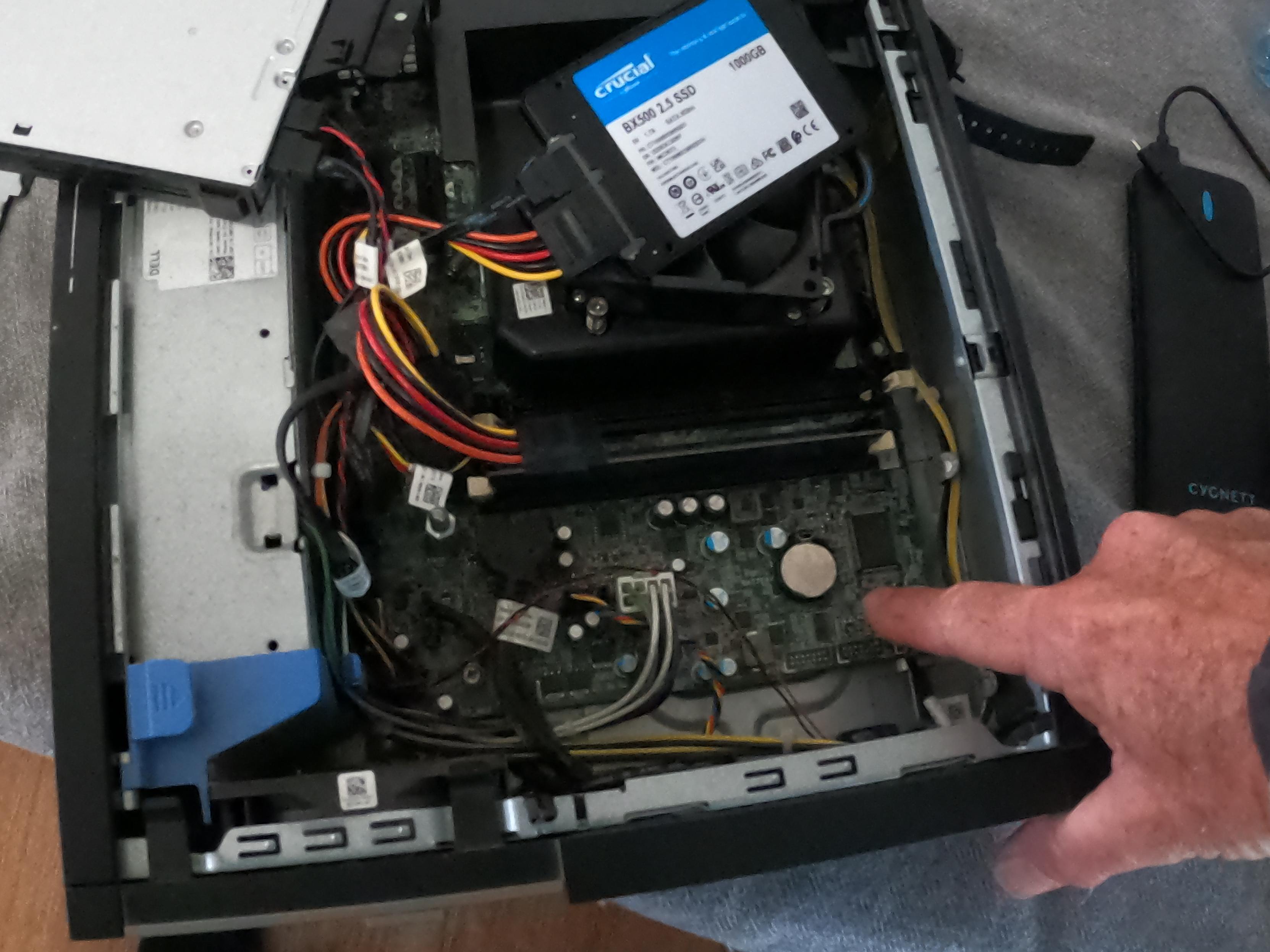 A PC with the lid off, a finger points to the button cell battery on the motherboard.