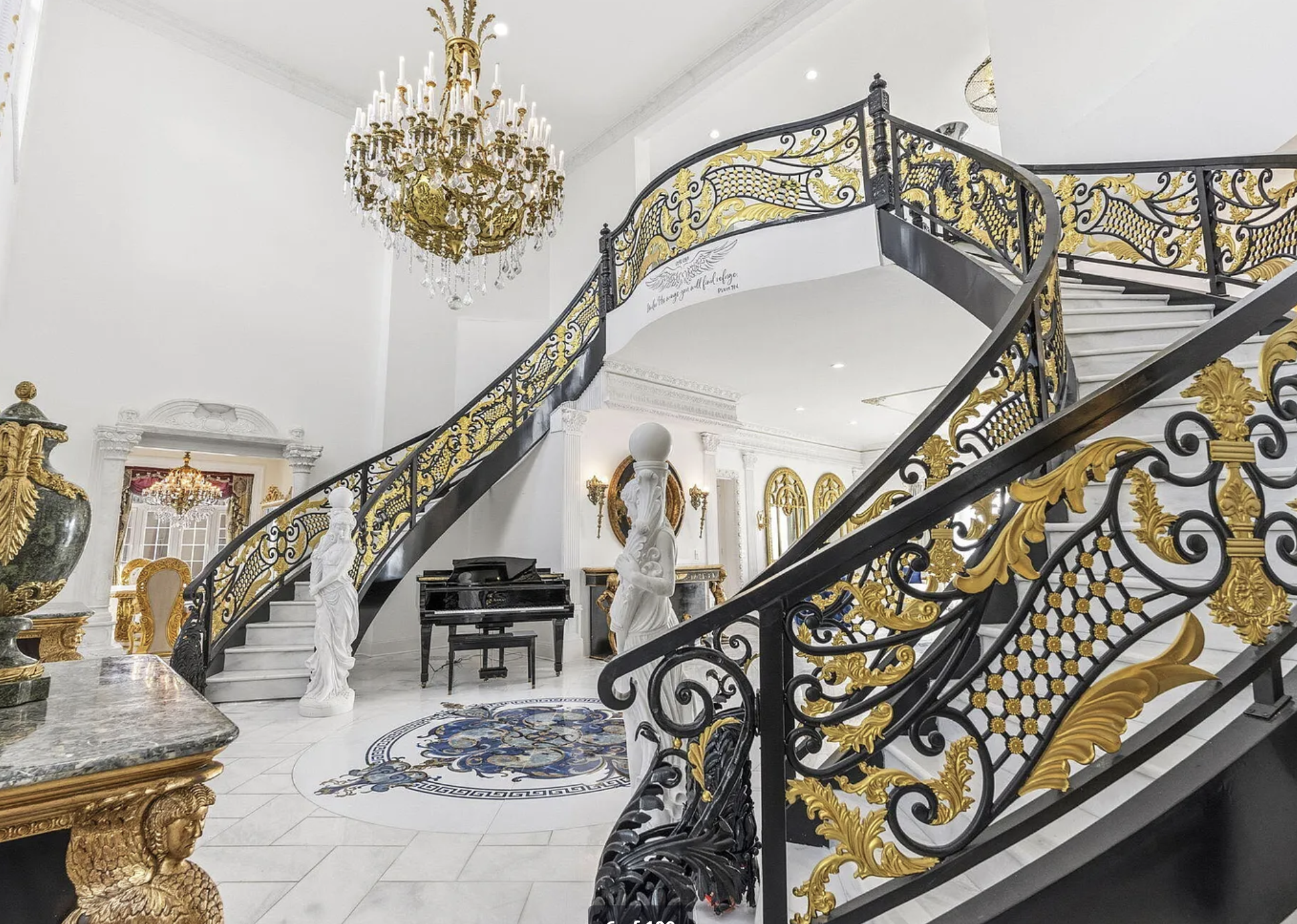 A foyer with a sweeping double staircase that is painted black and gold, and the floor is marble and there is a gold chandelier and gold furniture.
