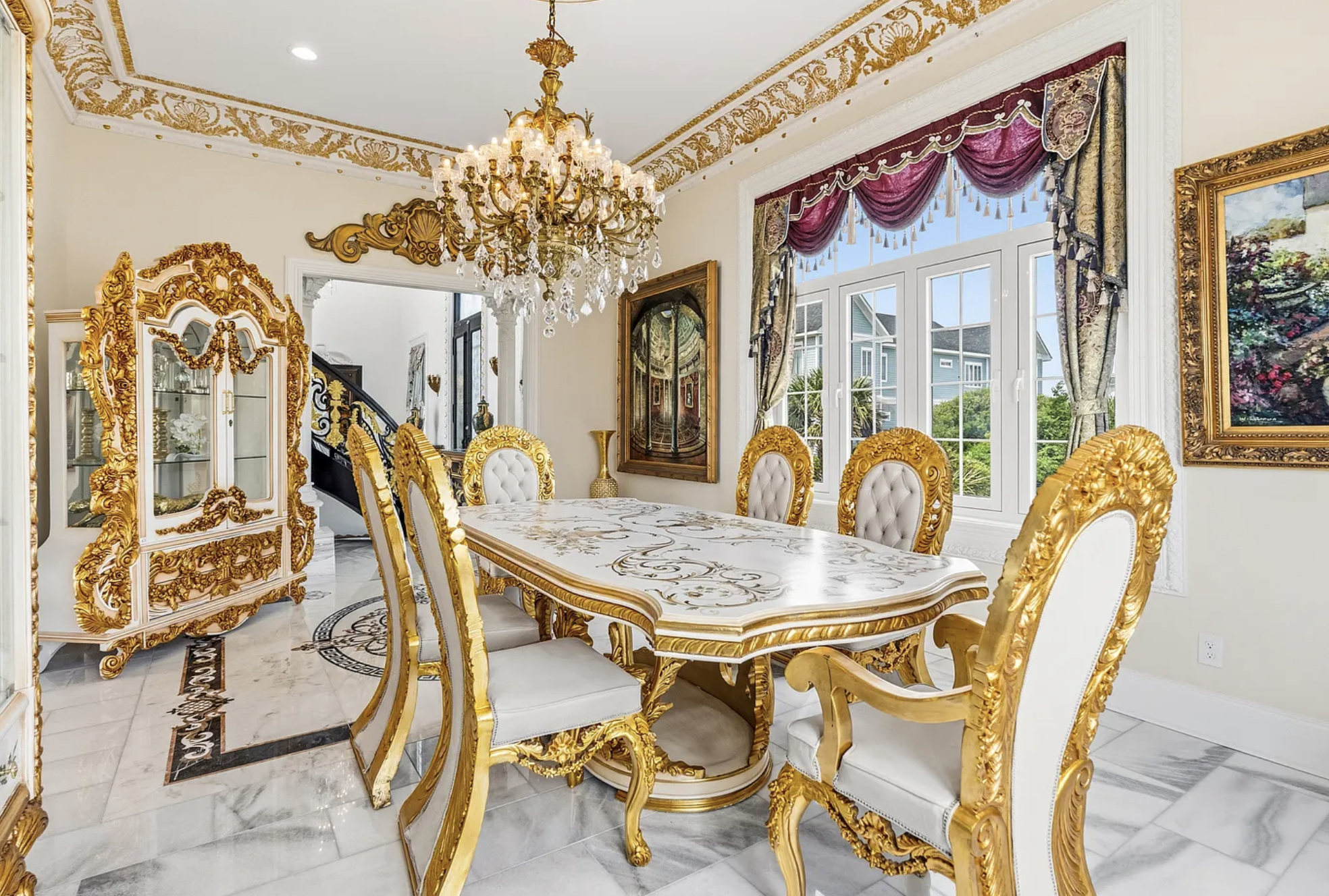 A dining room with gold painted trim around the ceiling, a large gold chandelier and a table and cabinet also painted white and gold.