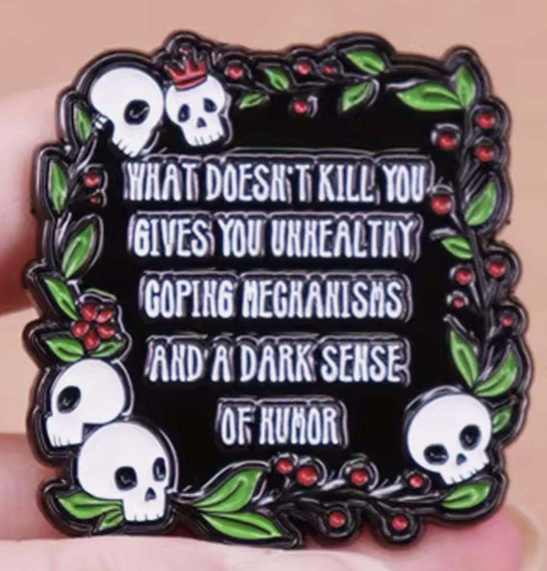 A pin Brooch decorate with skulls, wreath and the text “What doesn’t kill you gives you unhealthy coping mechanisms and a dark sense of humor”
