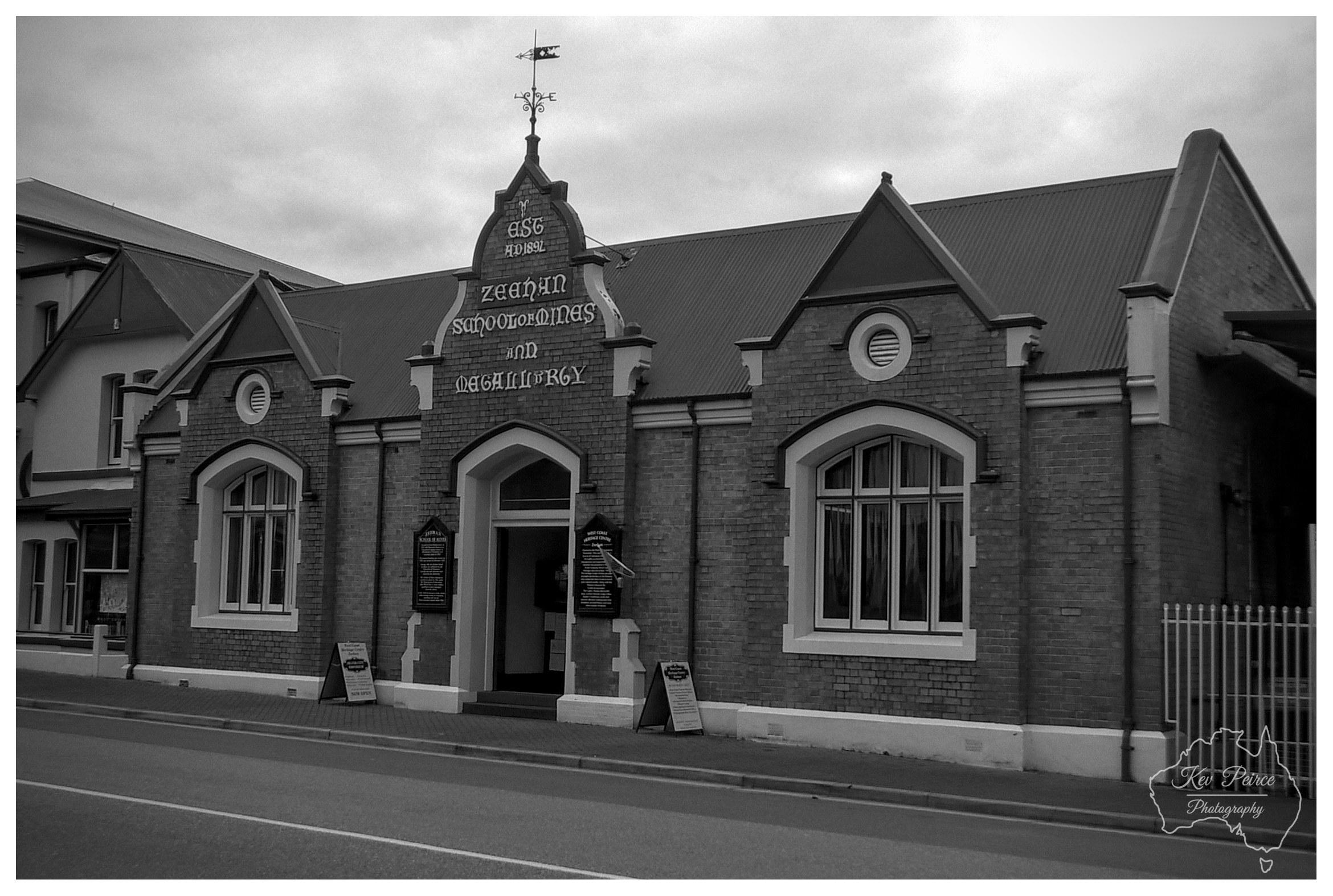 A monochrome, eye level photograph capturing the historic Zeehan School of Mines and Metallurgy building. The single storey brick structure features decorative gables, white stone trim, large framed windows, and a prominent central entryway topped with a weather vane and decorative lettering reading "EST AD 1892". The scene is captured under an overcast sky, casting a soft, even light across the textured brick facade and adjacent street.