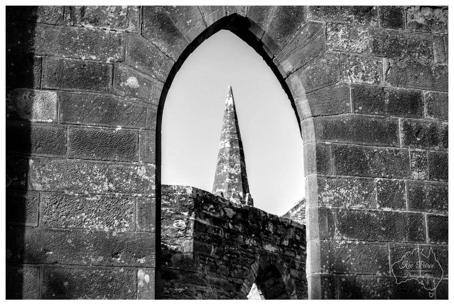 A high contrast black and white detail photo focused tightly through a pointed gothic archway made of large stone blocks. Positioned perfectly in the center of the arch in the distance is a sharp, pointed stone spire rising above ruined stone walls against a pale, cloudless sky.