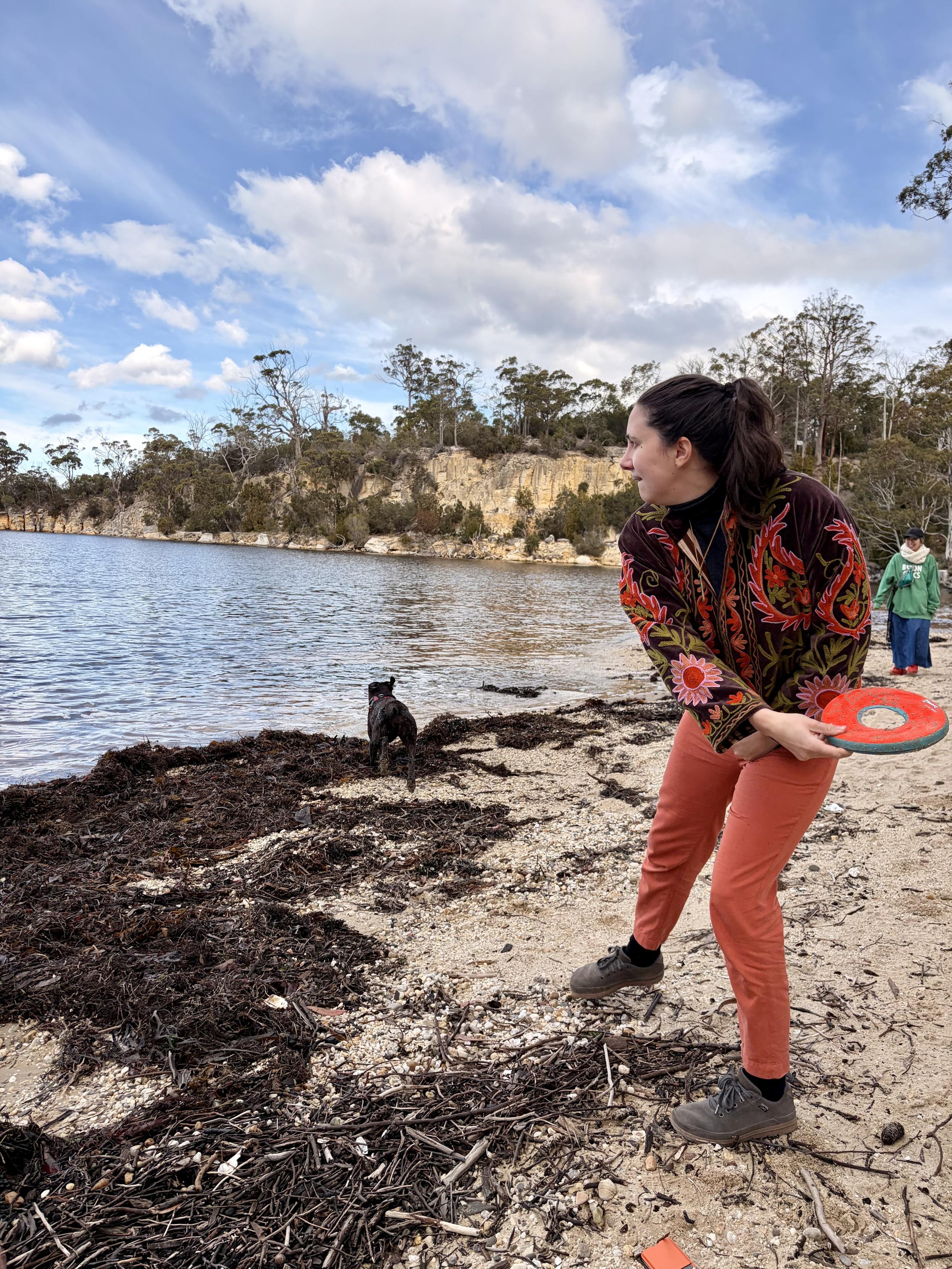 A friend. She has orange pants on with a colourful jacket. She is a beach with a dark brown dog in background. She has a bright orange frisbee throwing it out into the sea.