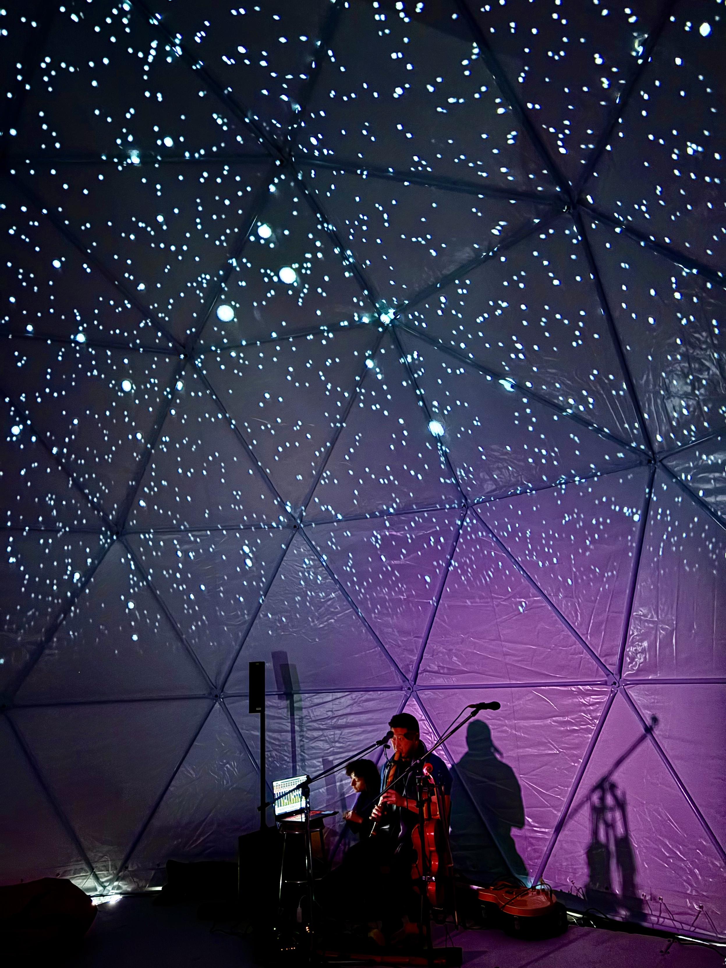 A man playing a flute in a dome. Lots of stars projected behind him with dark blue and pink.