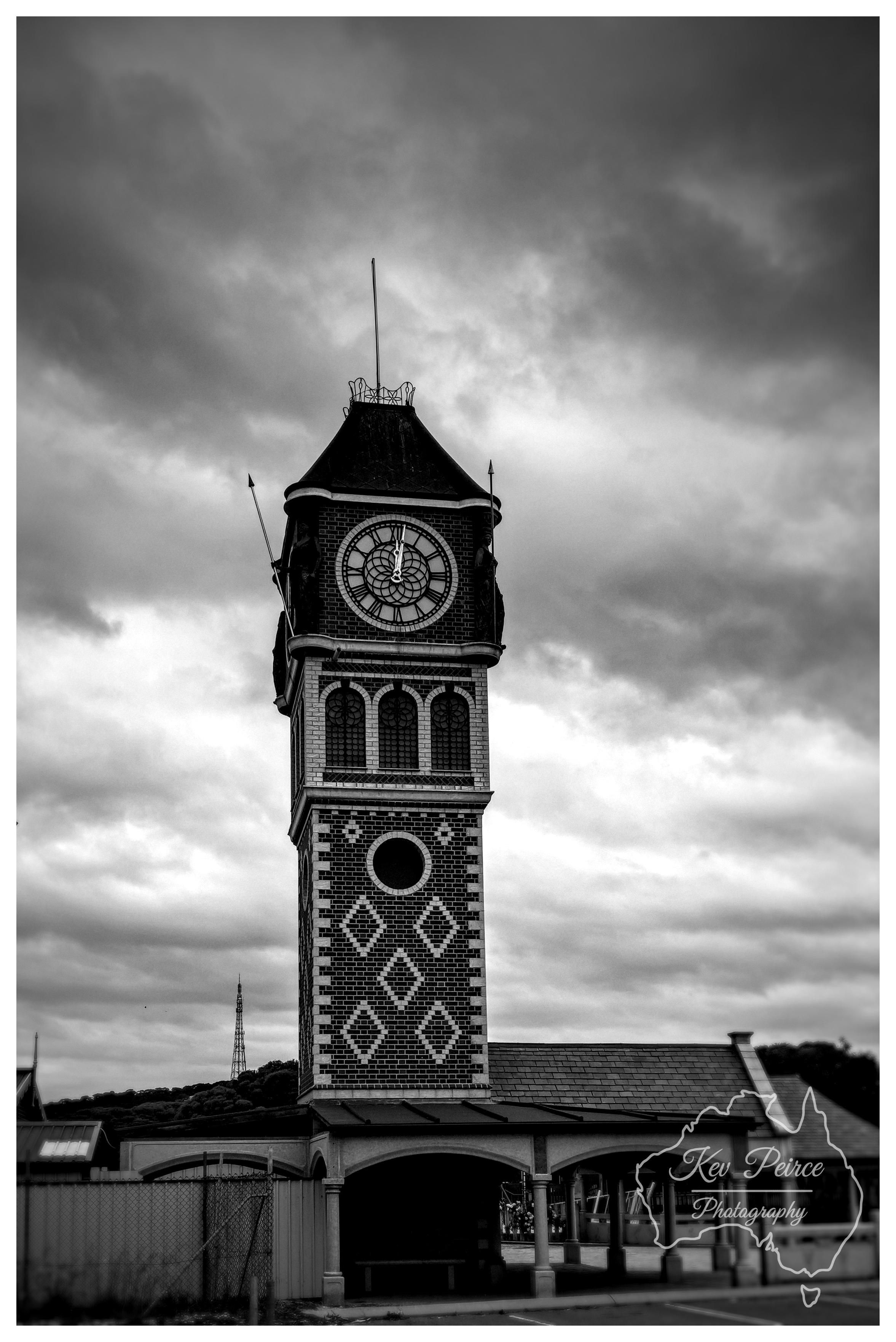 A vertical black and white photograph of an ornate, brick clock tower standing against a moody, heavily clouded sky. The tower features intricate brick patterning with white diamond motifs, arched upper windows, and a large central clock face near the top. Soft, diffused lighting accentuates the dark contrast of the bricks and the drama of the overcast sky. A low roofed structure with stone arches extends from the base of the tower.