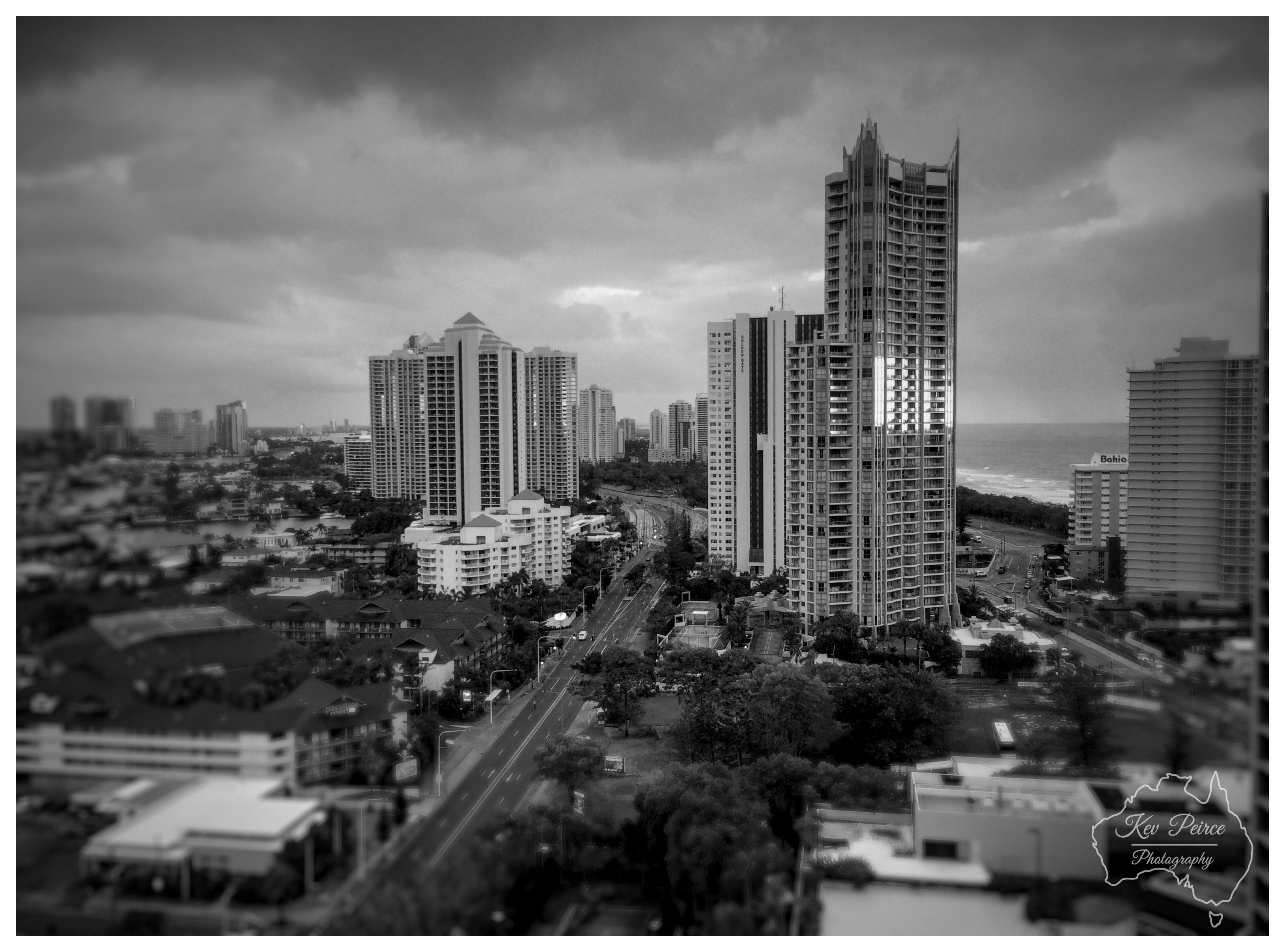 A monochrome aerial photo capturing a high rise coastal skyline under a moody, overcast sky. Sunlight breaks through the clouds, striking the glass facade of a prominent skyscraper and creating a brilliant highlight near the centre right. A main road cuts through lower buildings and lush trees in the foreground, stretching towards the distant horizon where the ocean meets the shore. The image features a strong tilt shift effect, with sharp focus across the central architecture and a soft blur fading into the top and bottom borders.