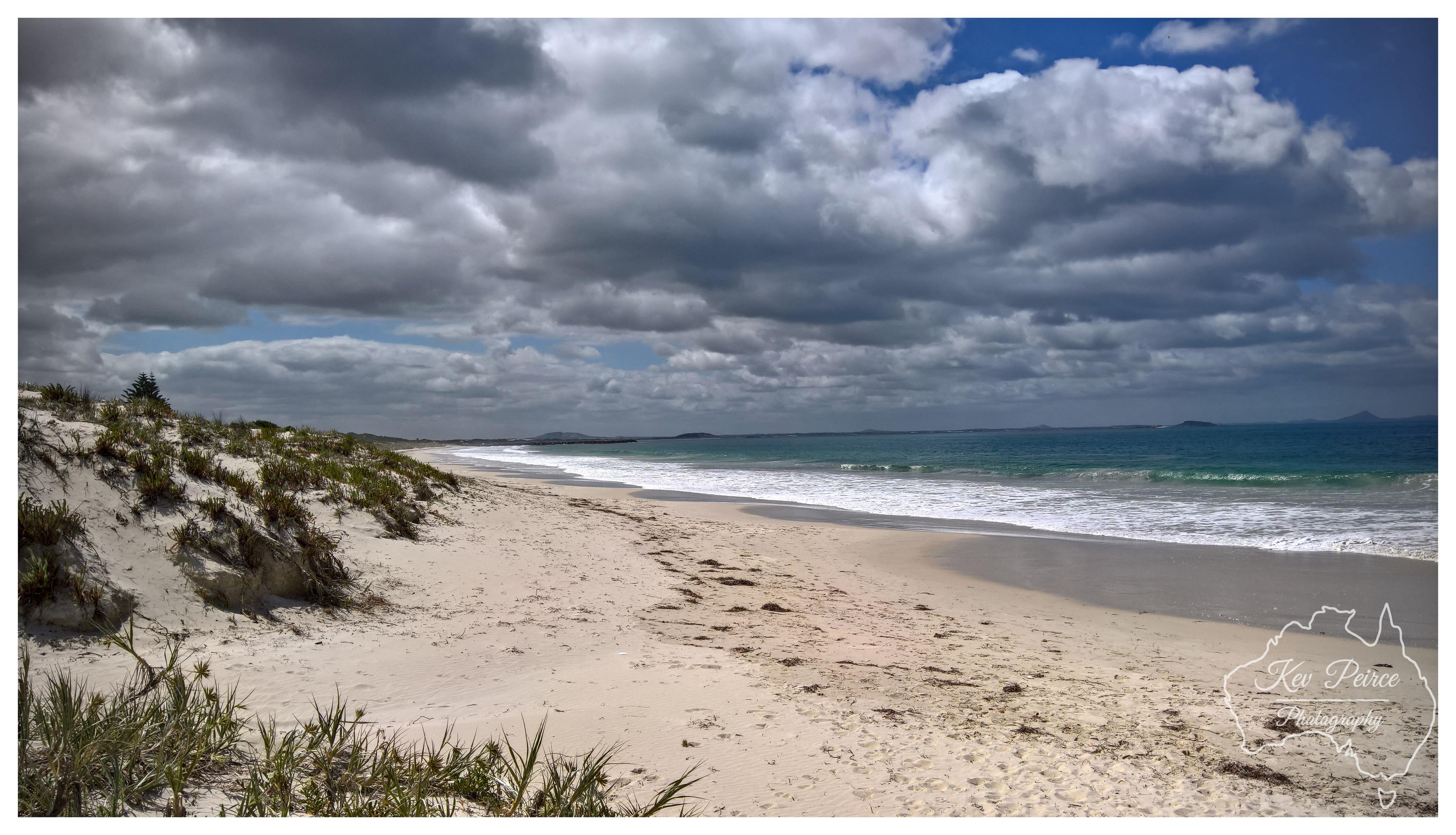 Wide angle view of a sweeping, empty sandy beach leading toward a turquoise sea. Coastal dunes covered in sparse green vegetation curve along the left side of the frame. Dramatic, textured grey clouds fill the sky above the water.