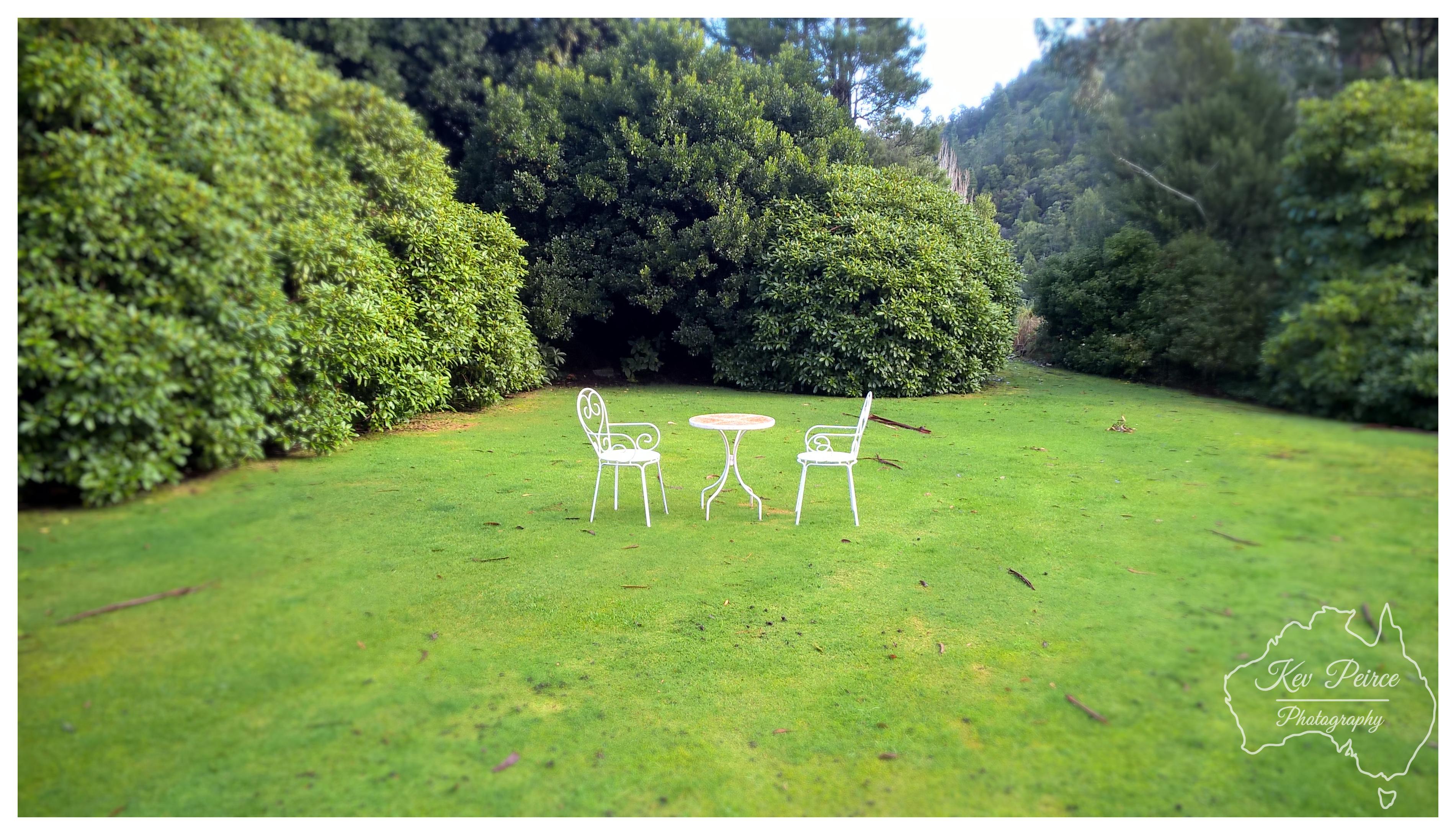 A wide shot of a neatly manicured green lawn surrounded by tall, dense green hedges and trees. In the middle of the lawn sits a decorative white outdoor setting, consisting of two metal chairs and a small round table. Overcast light creates an even, soft brightness across the entire garden space. A watermark map outline sits in the bottom right corner.
