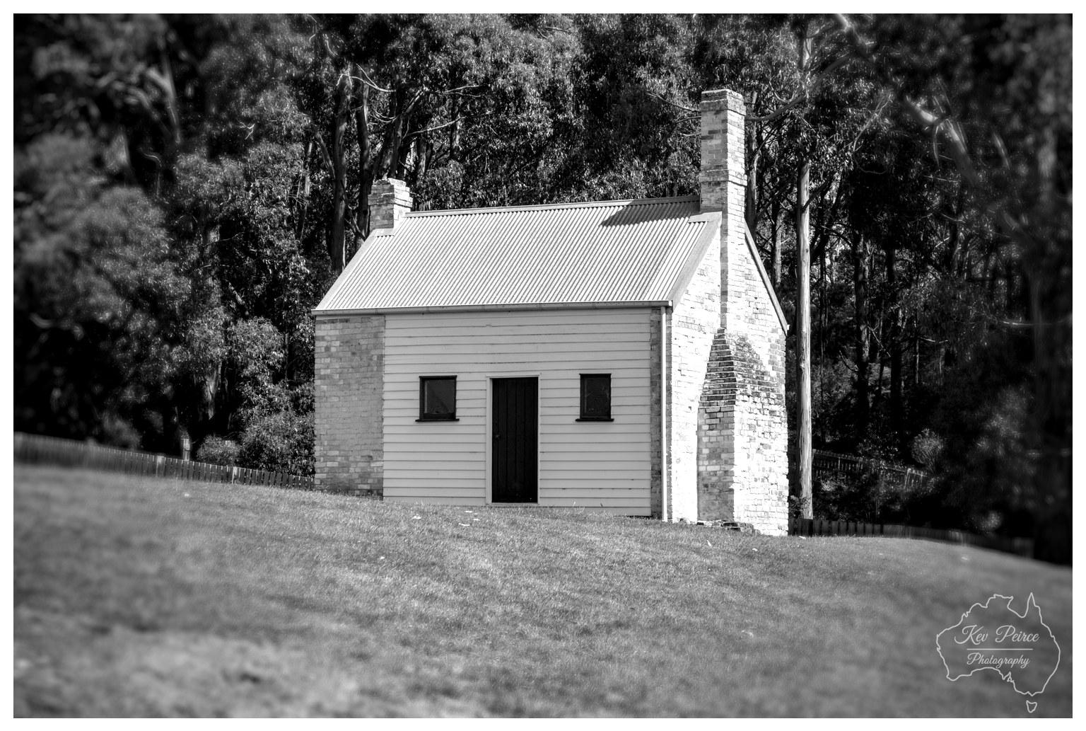 A monochrome image featuring a small, single storey timber cottage with weatherboard cladding and a corrugated iron roof situated on a sloping lawn. Two dark stone chimneys rise on either end of the cottage. High-contrast sunlight highlights the front facade, while dense, dark eucalypt trees create a heavily textured background behind the cottage.