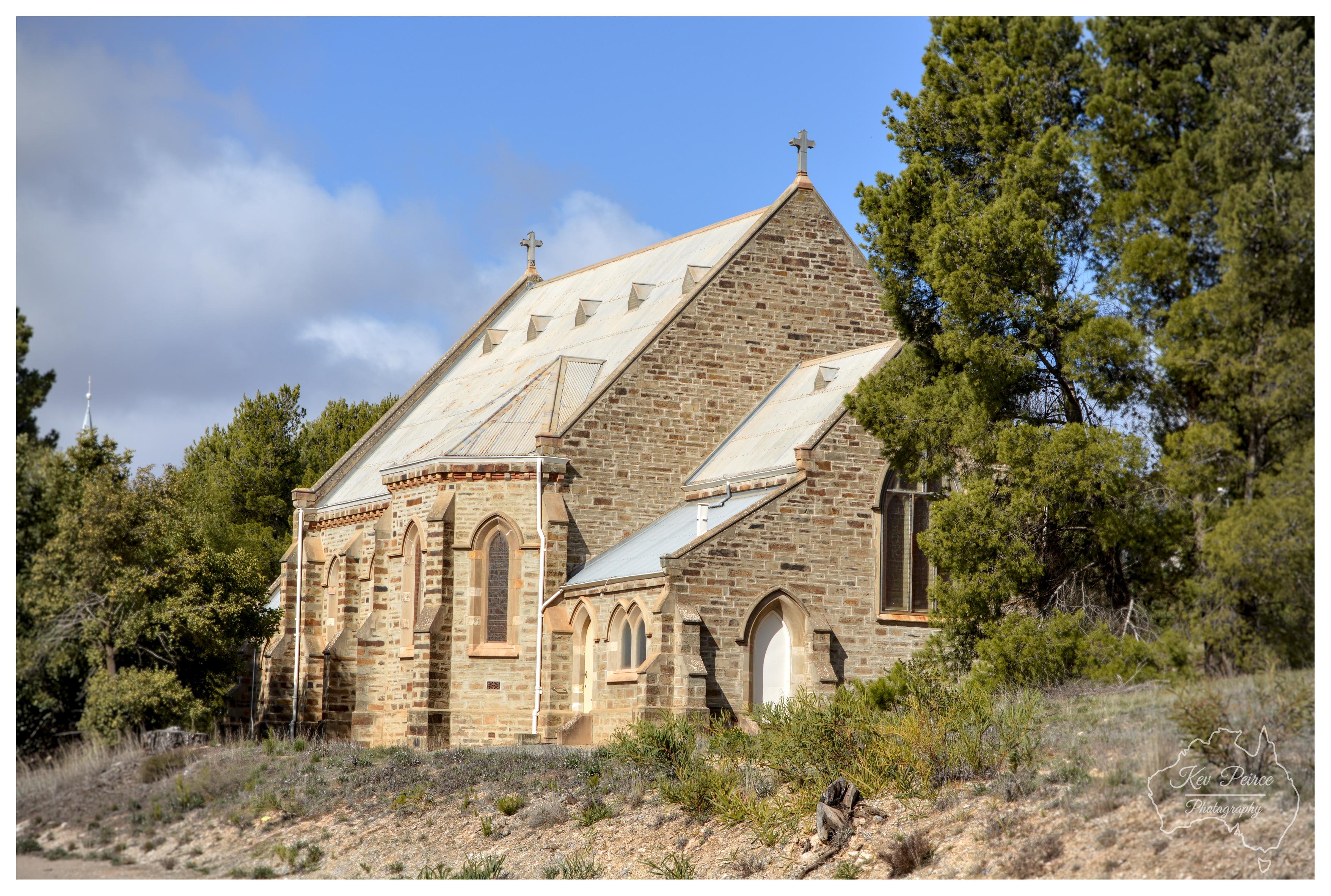 An historic stone church with Gothic Revival architecture, featuring pointed arch windows and two crosses on the gabled roof peaks. The weathered brick building sits on a gentle slope surrounded by native trees and shrubs under a blue sky with scattered clouds. Photo credit: Kev Peirce Photography.
