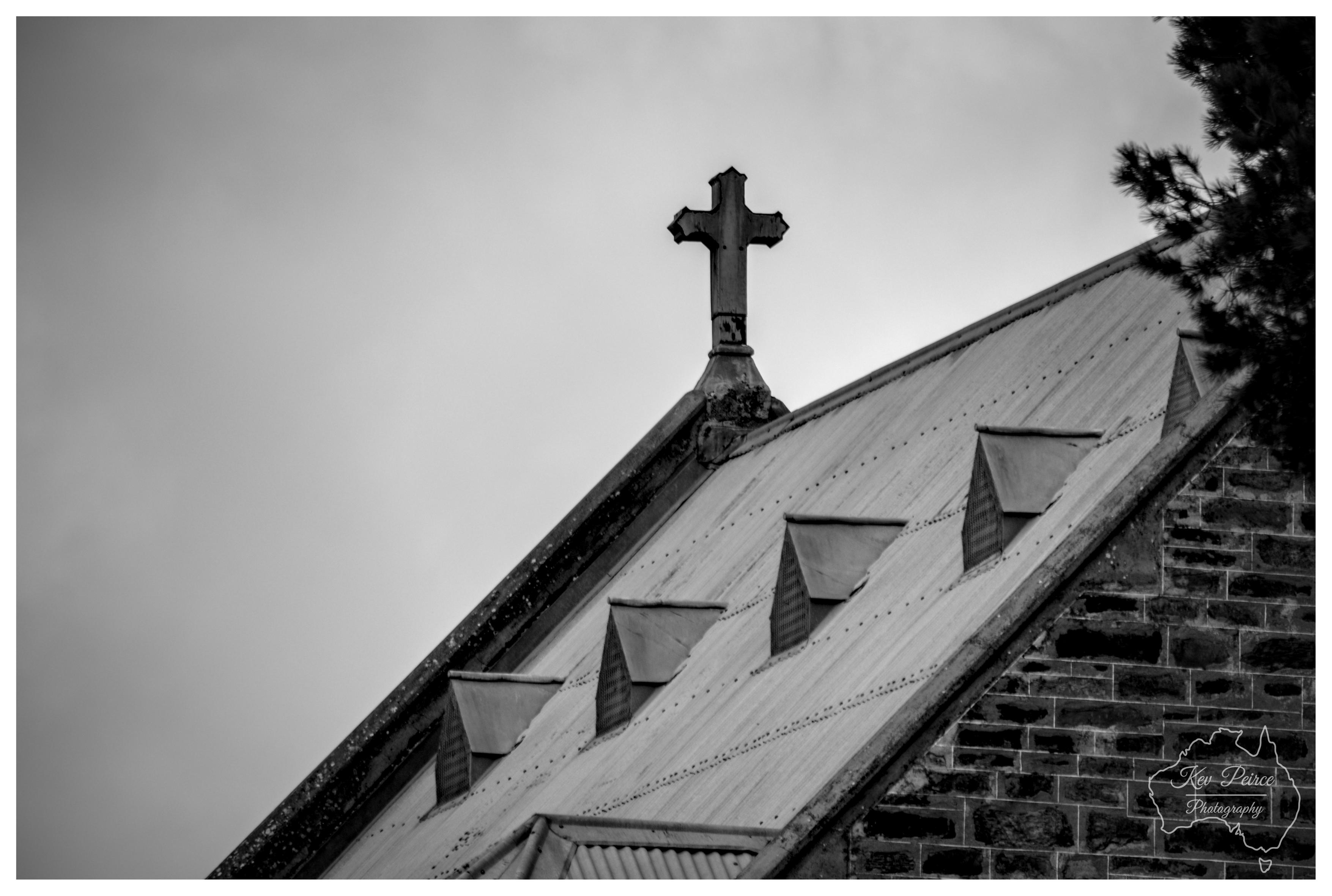 Black and white close up photograph of a weathered church cross mounted on the peak of a corrugated metal gabled roof. Small dormer vents line the sloping roof surface below, with stone masonry visible at the right edge and pine tree branches in the corner.