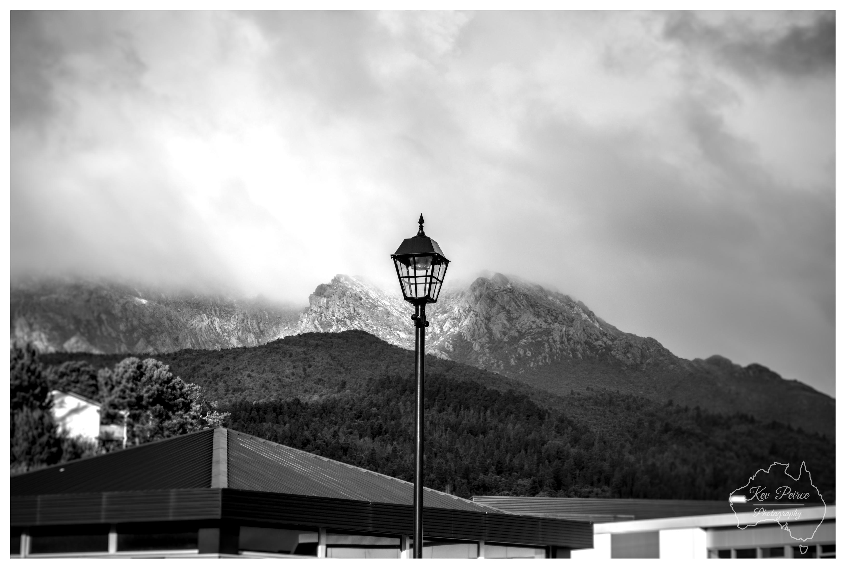 A black and white photograph featuring a traditional black street lantern in the center foreground. Behind the lantern, dark forested hills lead up to rugged, rocky mountain peaks partially shrouded in thick, overcast clouds. In the lower left, the dark roofline of a modern building provides structure to the scene. A watermark map logo rests in the bottom right corner.