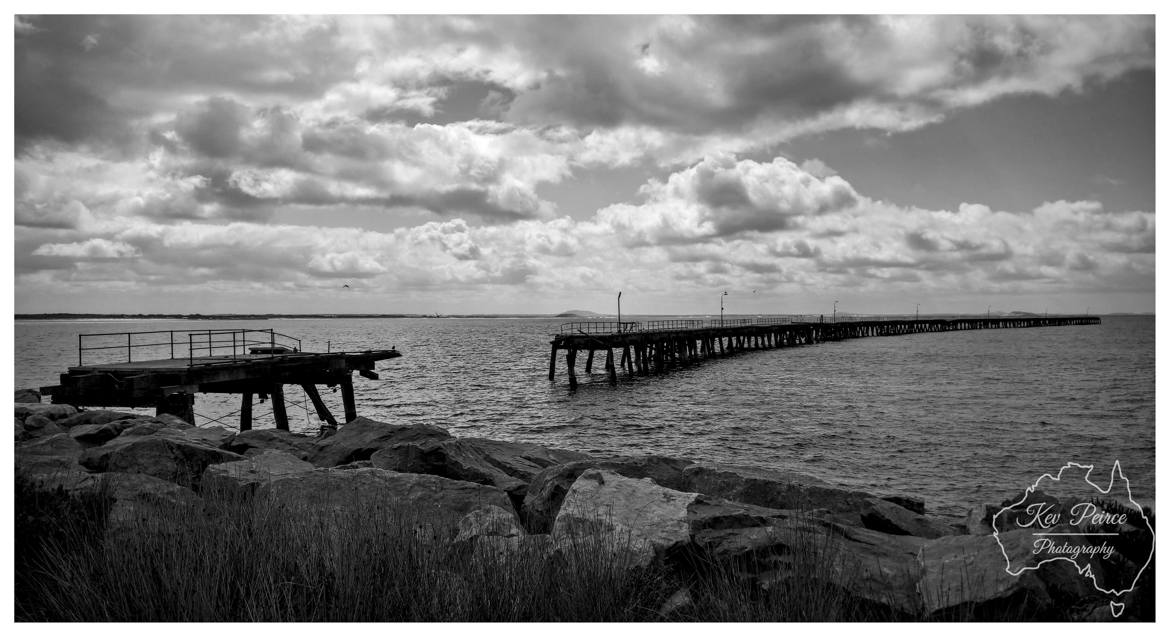 Black and white panoramic photograph of two wooden piers extending into calm water from a rocky shoreline. The longer pier stretches far into the distance while a shorter broken section sits closer to shore. Dramatic cloud formations fill the sky above. Water ripples gently between the structures. Bottom right features a signature watermark "Kev Peirce Photography" in an Australia shaped outline.