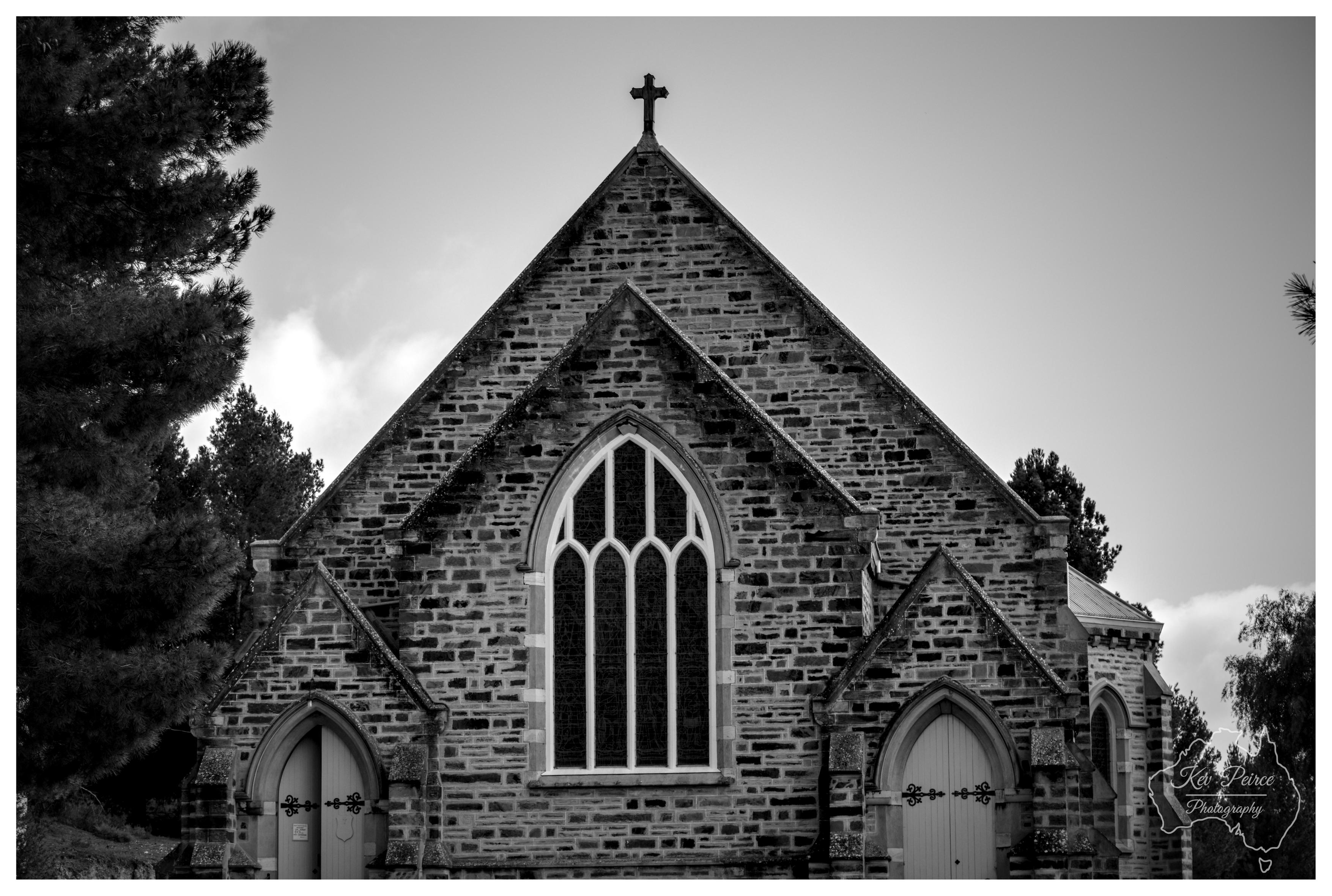 Black and white front view of a historic stone church featuring Gothic Revival architecture. Large pointed arched stained glass window dominates the centre, flanked by two smaller arched doorways with decorative iron hardware. A cross crowns the main gable peak. Pine trees frame the left side under dramatic sky.