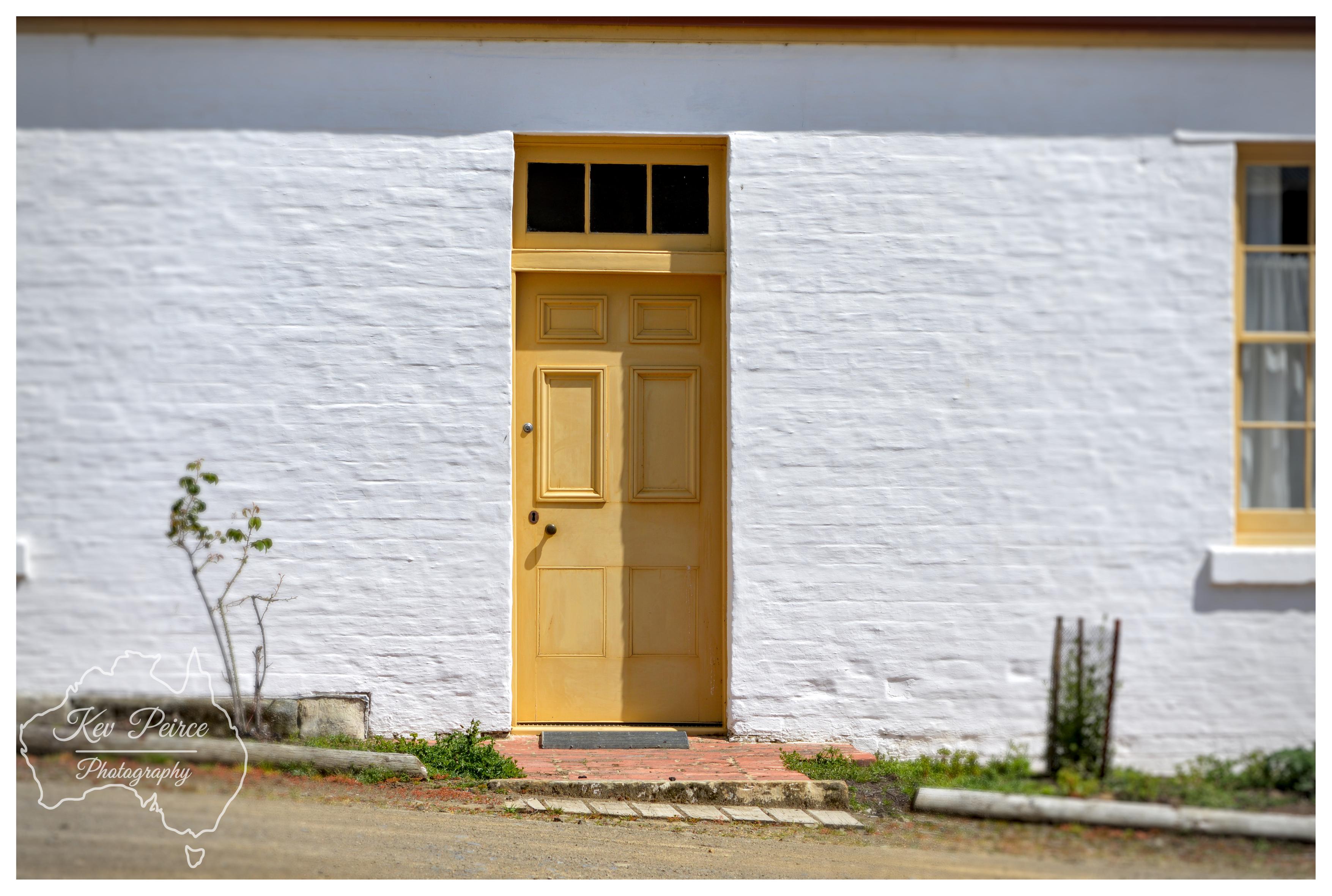 A clean, eye level architectural shot focusing on a pale yellow timber front door framed within a bright, white washed brick exterior wall. Warm sunlight highlights the door, small brick doorstep, and window on the right. A small green plant grows alongside a dirt pathway in the lower foreground.