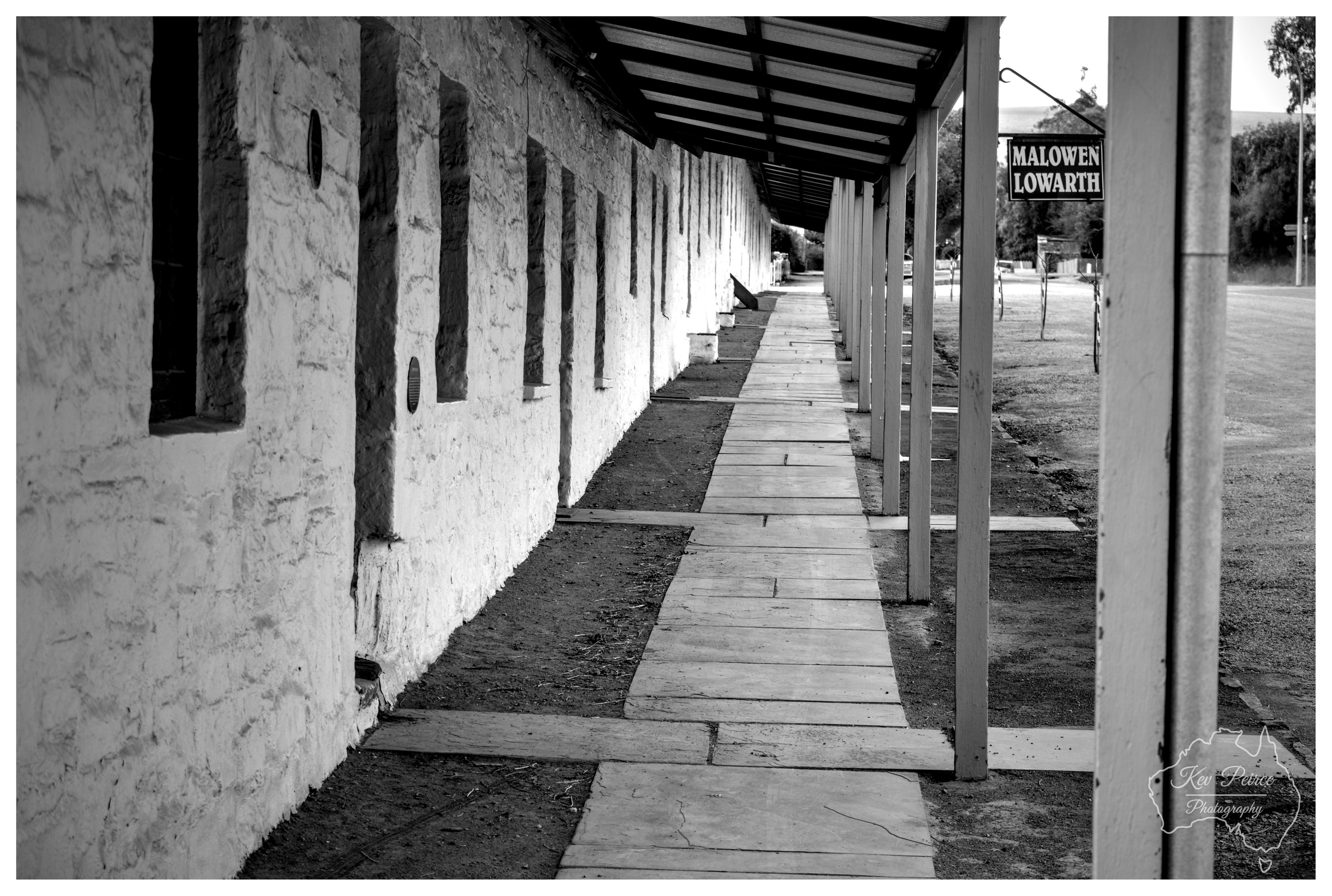 Black and white perspective shot of a historic covered walkway. Whitewashed rough stone wall runs along the left with evenly spaced rectangular windows. Wooden posts support a corrugated metal roof above a stone slab pathway. A hanging sign reading 'MALOWEN LOWARTH' is visible on the right, creating leading lines toward the vanishing point.