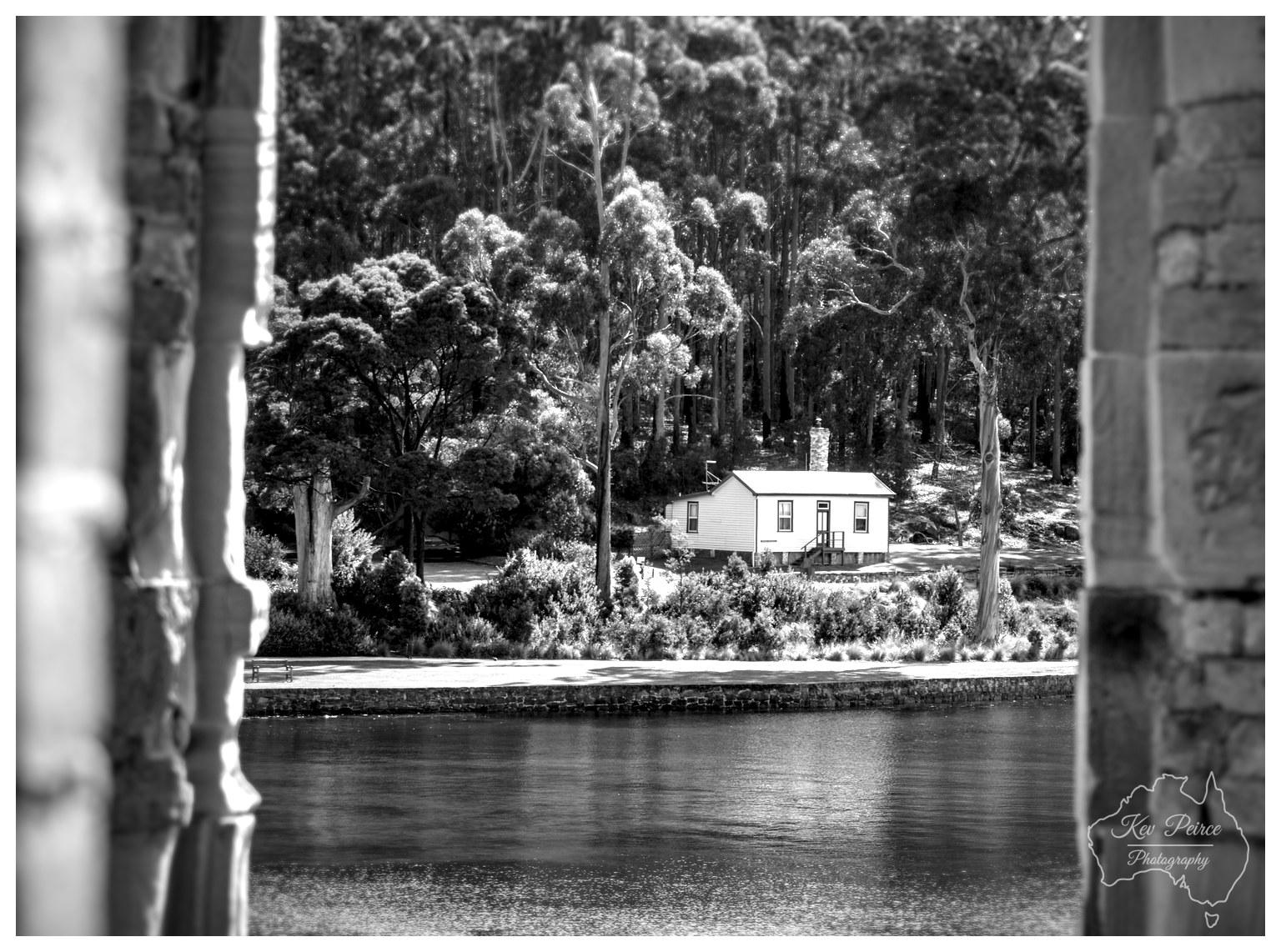 A black and white photograph framed by the thick vertical stone masonry of an adjacent archway in the foreground. Looking across a smooth body of water, a small white single story cottage sits on the far bank against a dense forest of tall trees. Sunlight reflects softly off the surface of the water, drawing the eye toward the distant home.