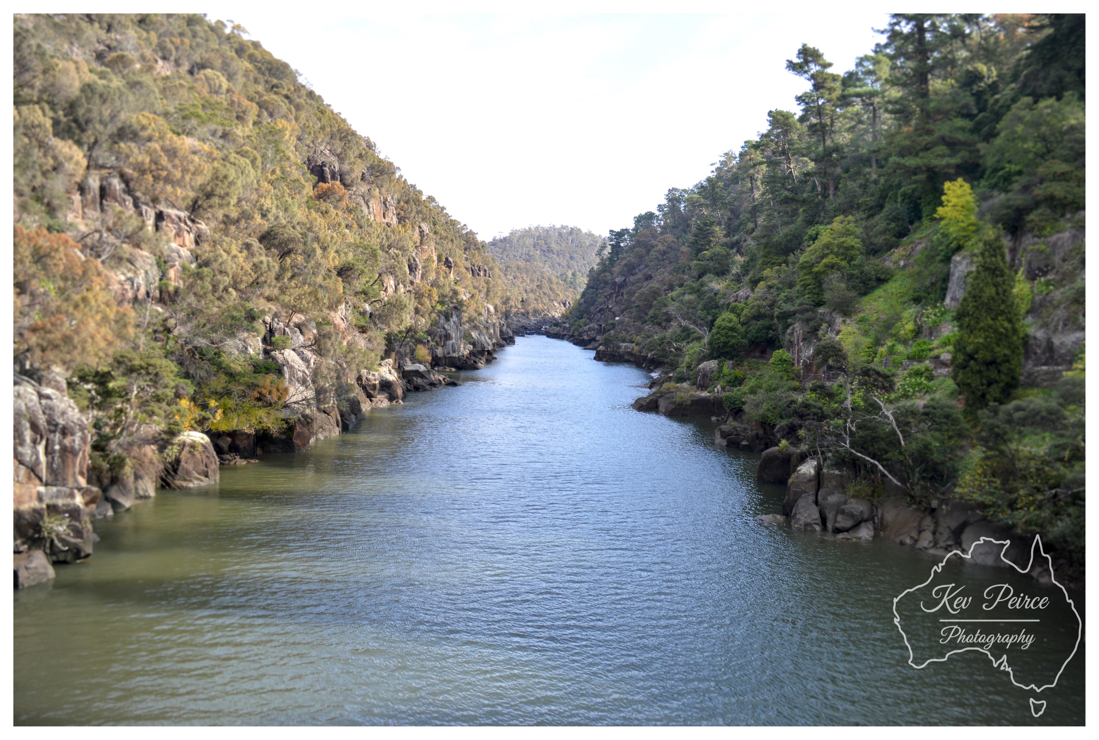 A wide landscape photograph of Cataract Gorge. A calm, wide river flows down the centre towards the distance, flanked by steep rocky cliff faces. The left bank features sunlit rocky slopes dotted with dry, olive toned native scrub, while the right bank is densely covered with lush green eucalypts and conifers. Soft, bright daylight illuminates the river surface, which reflects the pale sky above. In the bottom right corner, a white line art watermark outlines Australia with cursive photographer text.