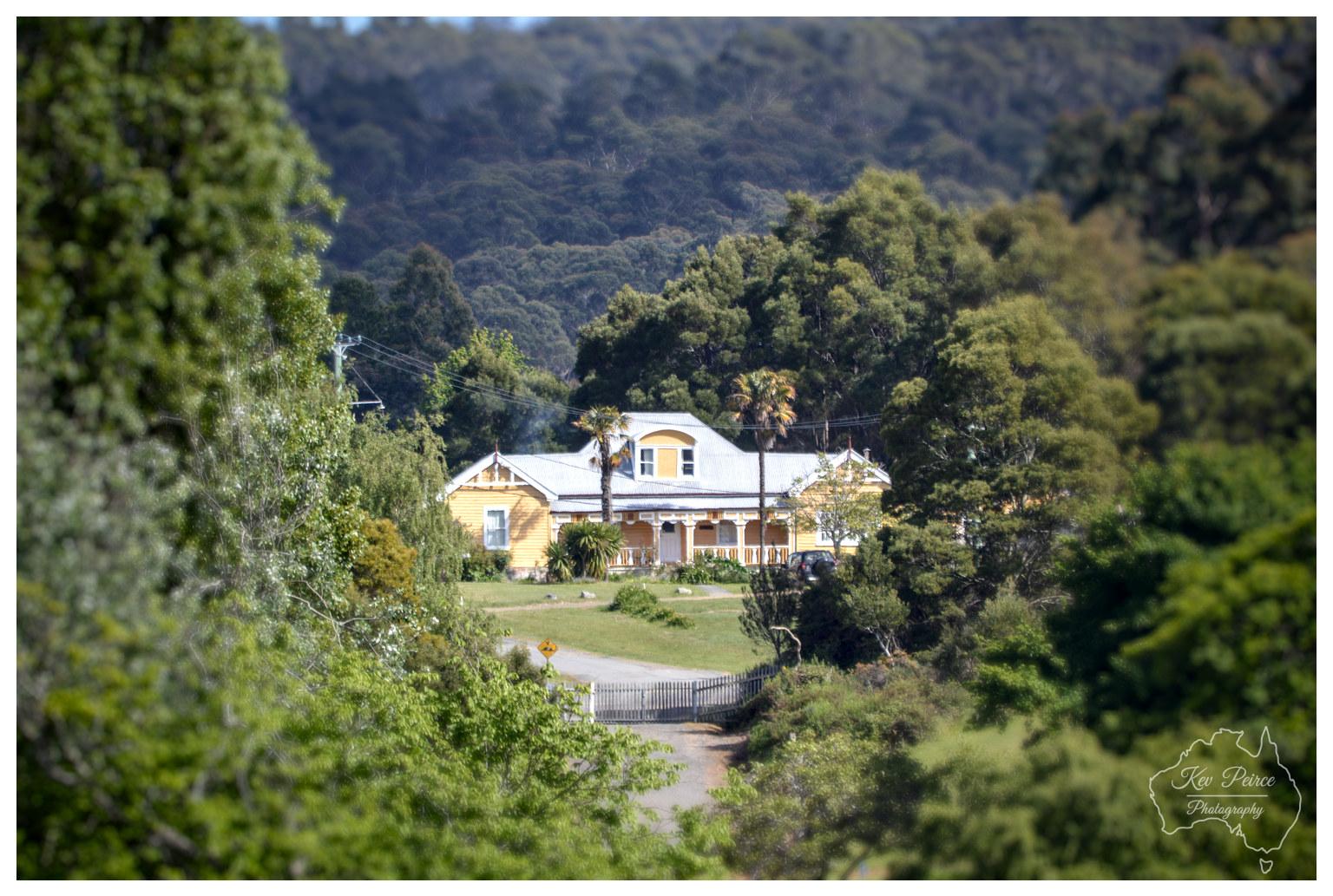 A telephoto shot looking down a narrow dirt lane framed by soft-focus green foliage in the foreground. A large two storey yellow timber cottage with white trimmed verandas and twin palm trees sits at the end of the track. Thick, rolling green hills covered in bushland rise high into the background under natural daylight.