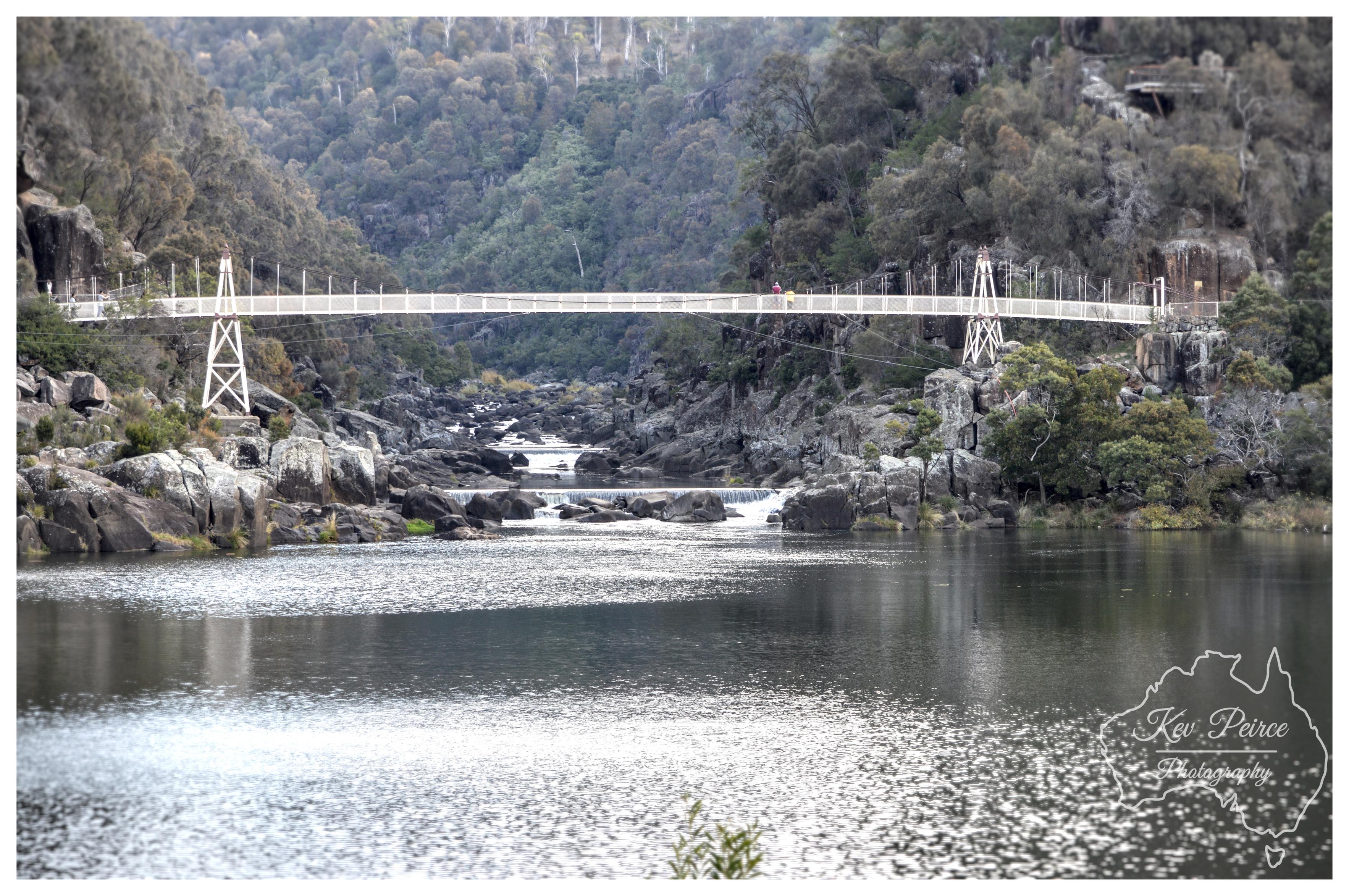 A wide landscape photograph featuring the white Alexandra Suspension Bridge stretching across Cataract Gorge. The foreground shows sparkling river water catching the daylight with a shimmering texture. Behind the open water, rugged granite boulders and small river rapids sit beneath the long, slender suspension bridge supported by white trestle towers on either bank. Distant figures of people can be seen crossing the span. In the background, steep hillsides rise up, blanketed in native grey-green forest and rocky outcrops. A white outline watermark of Australia sits in the lower right corner.