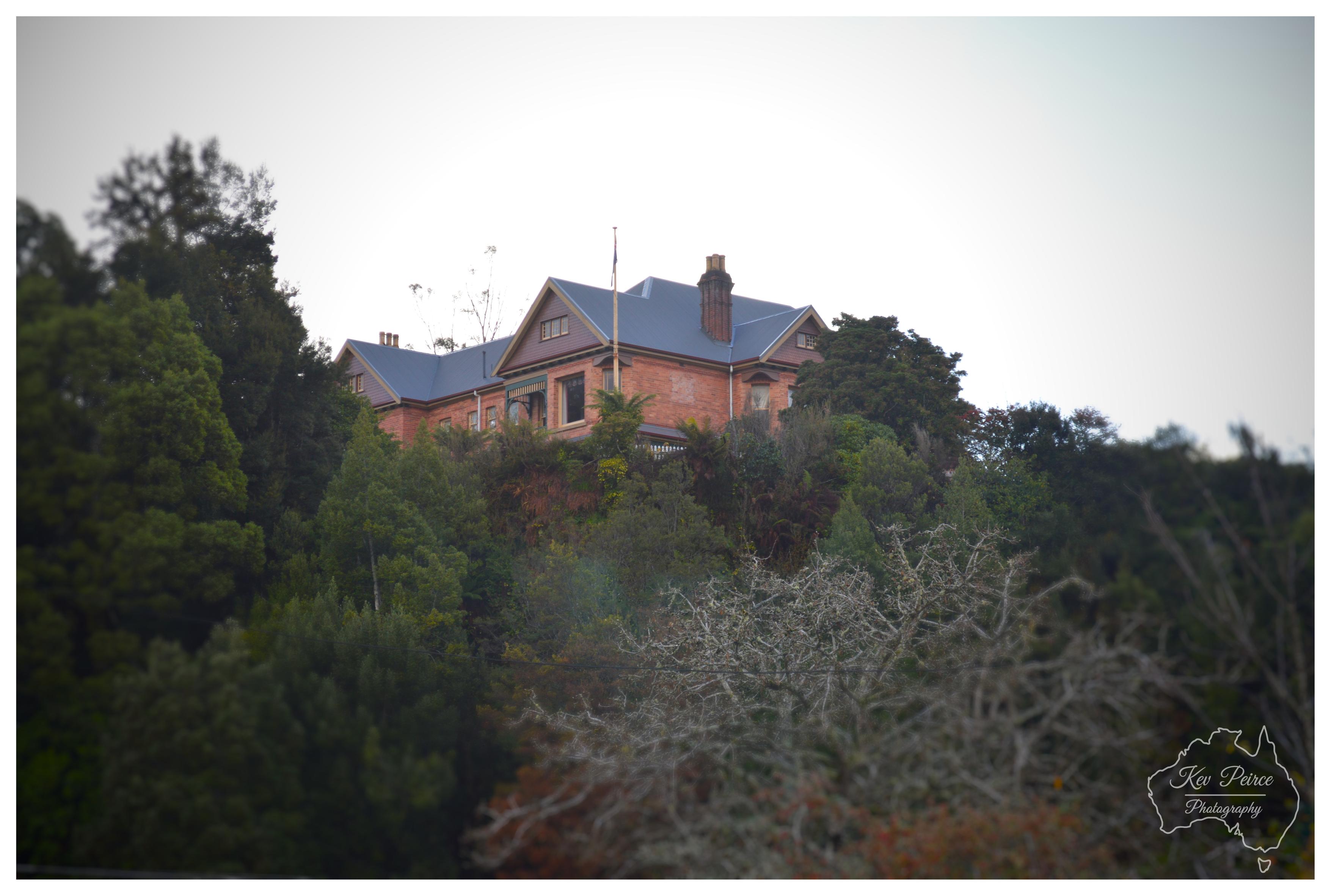A low angle shot showing a large, red brick historic manor house sitting atop a steep hill surrounded by dense, dark green trees. The building features multiple gables, chimney stacks, and dark blue roofing against a bright, overcast sky. The surrounding tree line spans the middle ground, while soft, out of focus foliage appears in the lower foreground. A subtle watermark map outline is present in the bottom right corner.
