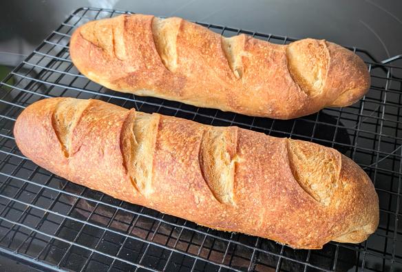 Two chubby little baguettes cooling on a wire rack. They have four score lines each and are a lovely burnished orange colour. The one at the rear is a little wonky on both the scoring and shaping, but it still looks delish.