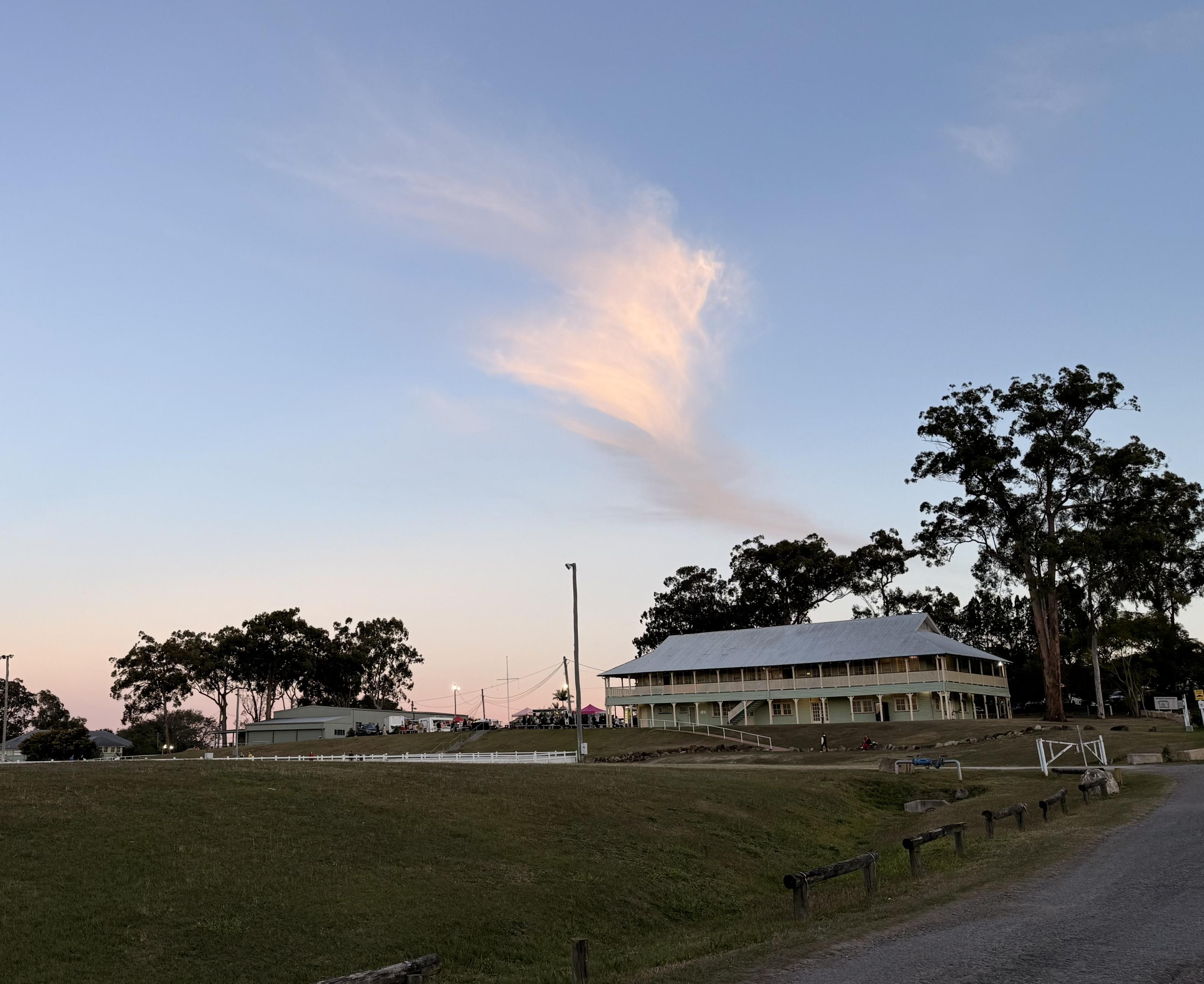 A sunset sky with a pink/orange cloud that looks almost like a soft arrow points to a two storey traditional Queenslander style wooden building, visible across a grassy field with a gravel road in front.