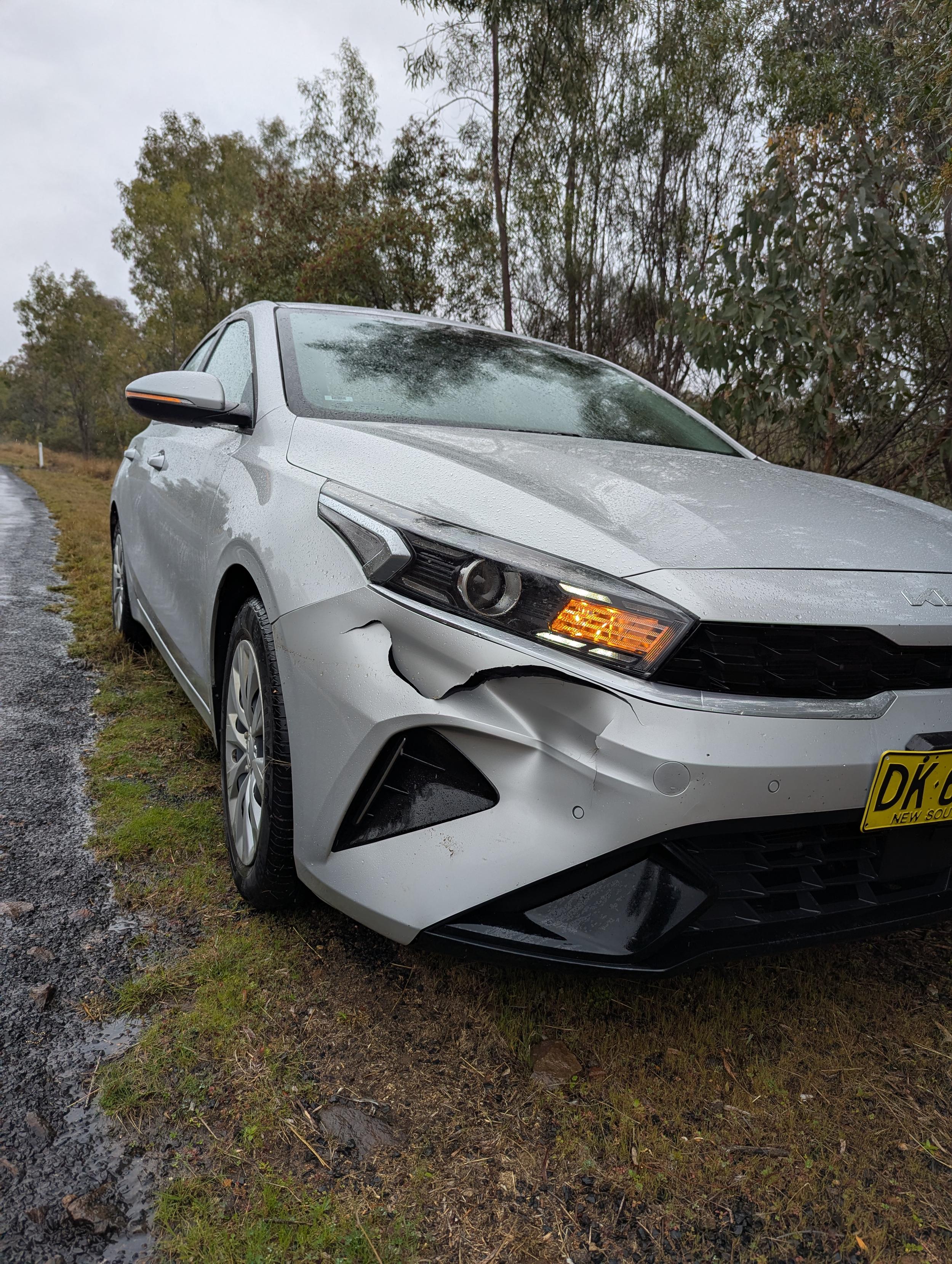 Photo of the front left corner of a car. The bumper is pushed in and cracked in several places from an impact.