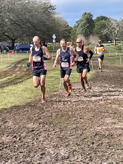 A group of runners in racing singlets and shorts run in a line along a muddy path.Trees and grass are visible in the background.