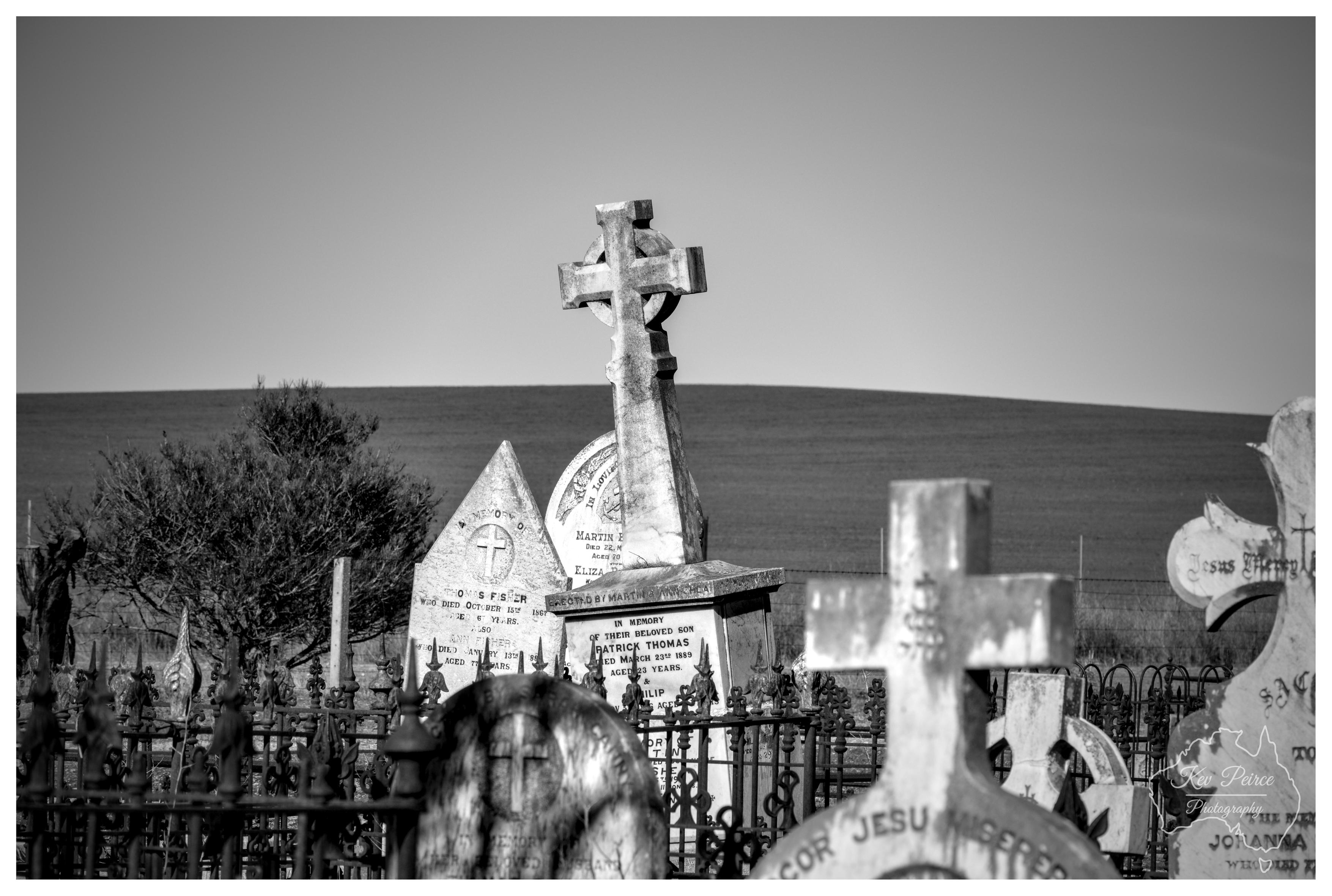 A monochrome photograph of an old rural cemetery surrounded by ornate wrought iron railings. A tall, weathered stone Celtic cross rises sharply in the centre, positioned alongside pointed and arched headstones carved with historic inscriptions. In the foreground, several aged cross headstones appear in a soft, shallow depth of field. A native shrub sits to the left, with the smooth, curved horizon of a low rural hill stretching across the background under a pale, clear sky in bright daytime light.