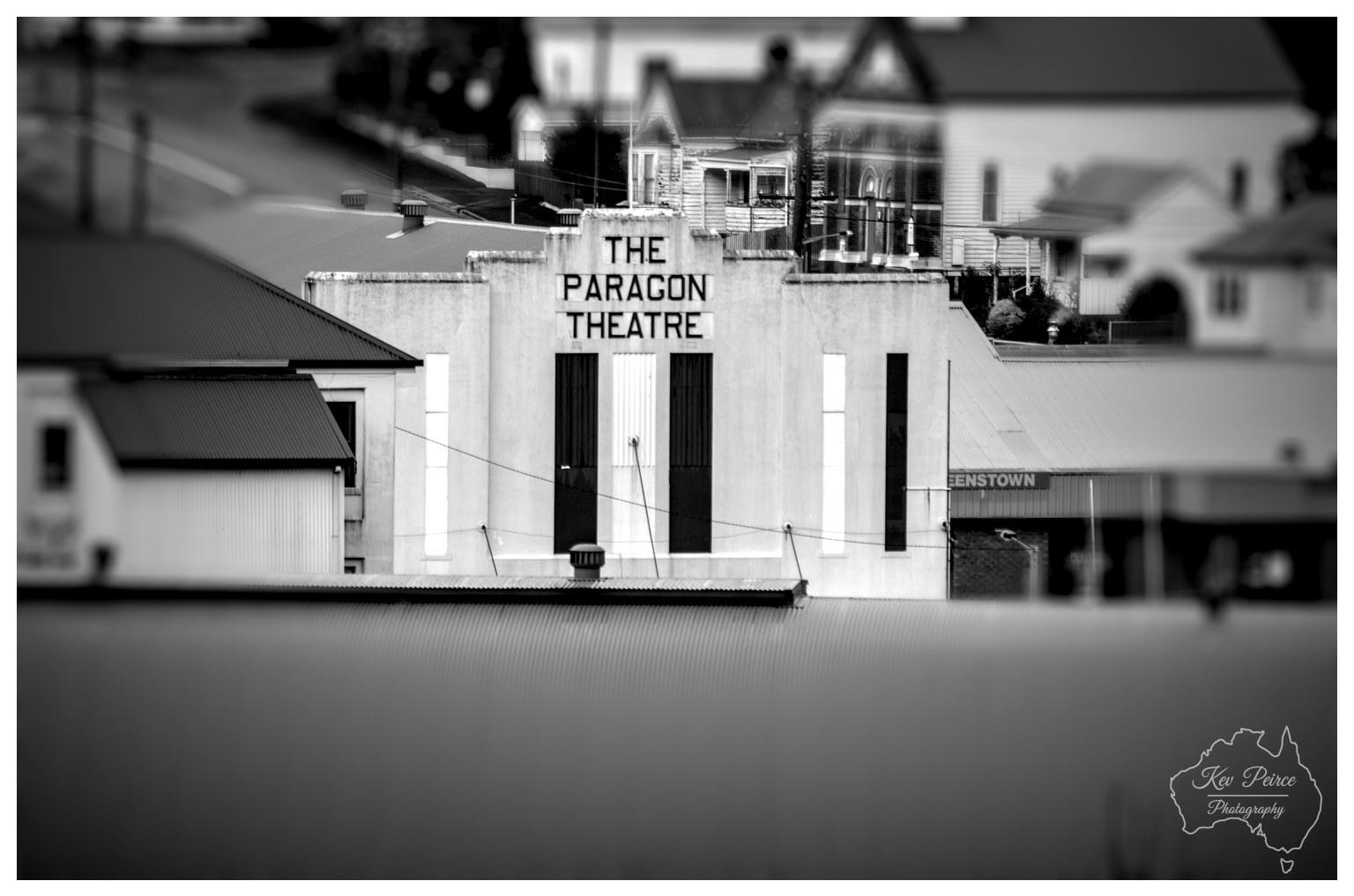 A monochrome, miniature effect photograph focusing sharply on the Art Deco facade of The Paragon Theatre in Queenstown. The building features bold vertical pillars, decorative fluting, and prominent dark lettering reading "THE PARAGON THEATRE" set against light stucco. The surrounding townscape, including corrugated iron rooftops, hilly streets, and regional weatherboard buildings in the background, is rendered in a soft, progressive blur, creating a tilt shift selective focus effect. A smooth, out of focus grey rooftop spans the bottom foreground, directing attention straight to the architectural details under soft, diffused daytime lighting.