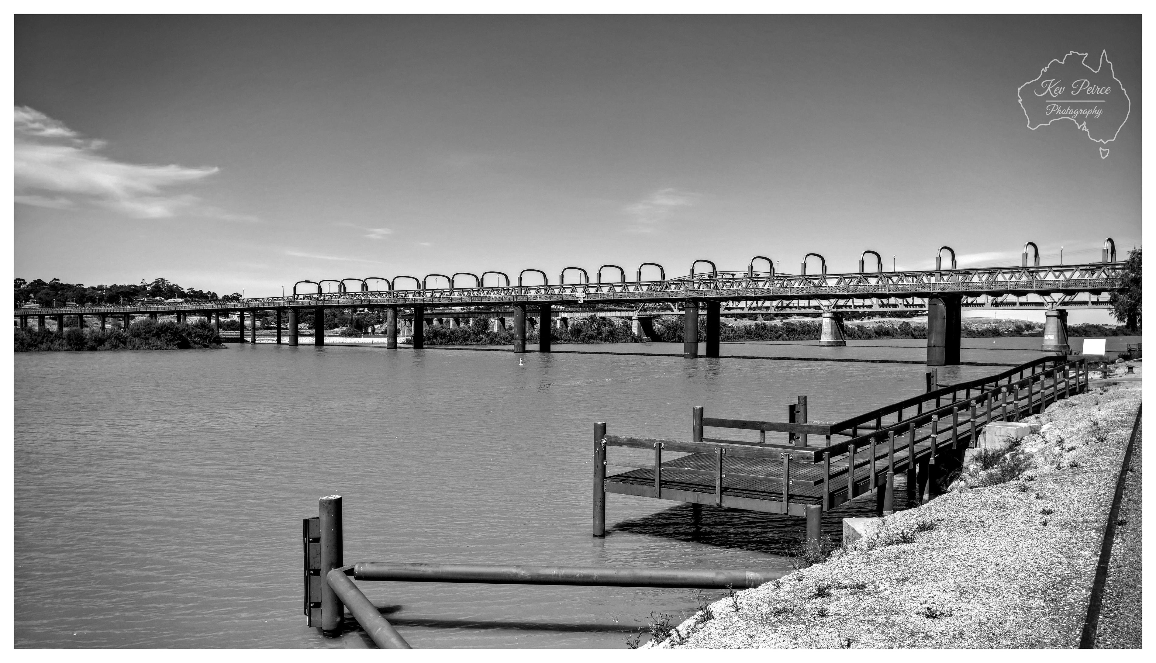 A wide angle monochrome photograph capturing the Murray Bridge road and rail spans crossing the calm waters of the Murray River. In the right foreground, a wooden jetty and walkway extend down a light, gravelly riverbank towards the water. The historic bridge stretches horizontally across the middle ground, showcasing rhythmic arched tubular trusses and sturdy pylons. Across the river, distant tree lined banks sit beneath a broad sky marked by wispy, light clouds under clear daytime illumination.