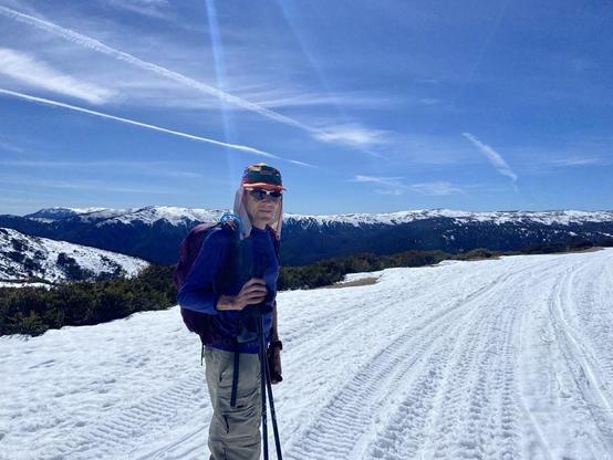 A man stands with ski poles on the snow, wearing a hat and sunglasses. A snow-covered mountain is behind them and the sky is blue with light clouds and contrails.