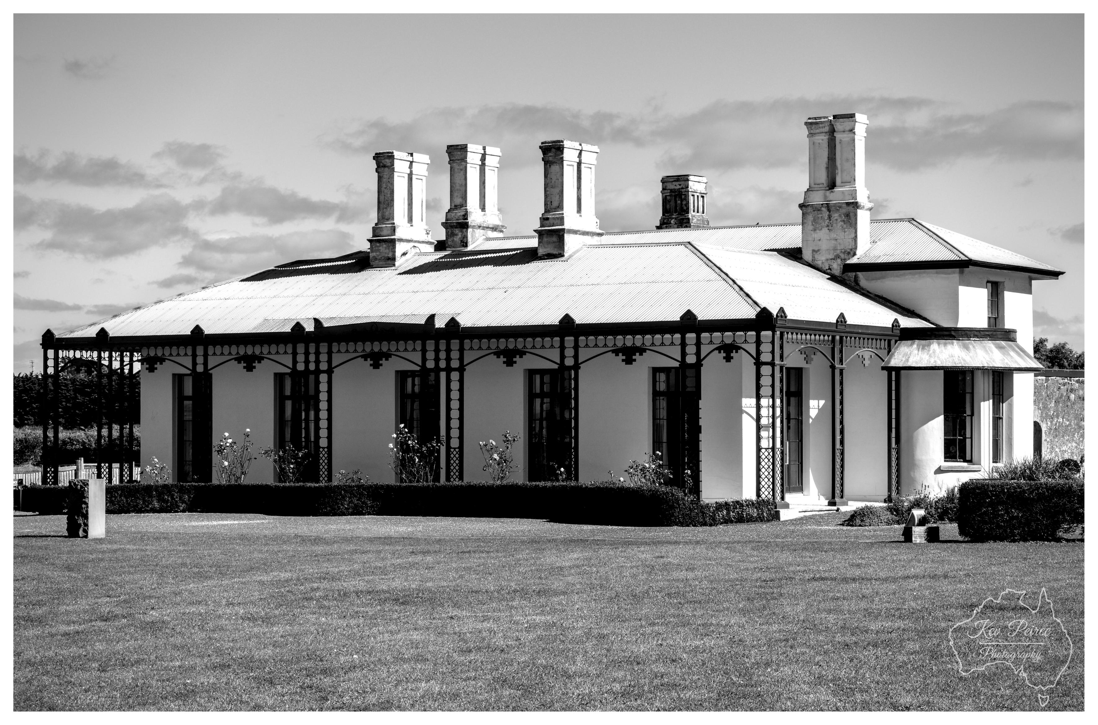 A wide monochrome landscape photograph capturing a grand colonial homestead set beyond a broad grassed lawn. The historic building features a corrugated iron hipped roof punctuated by five tall, decorative chimney stacks, an intricate ironwork verandah fringe, and shuttered French doors flanked by neat hedges and rose bushes under a lightly clouded sky.