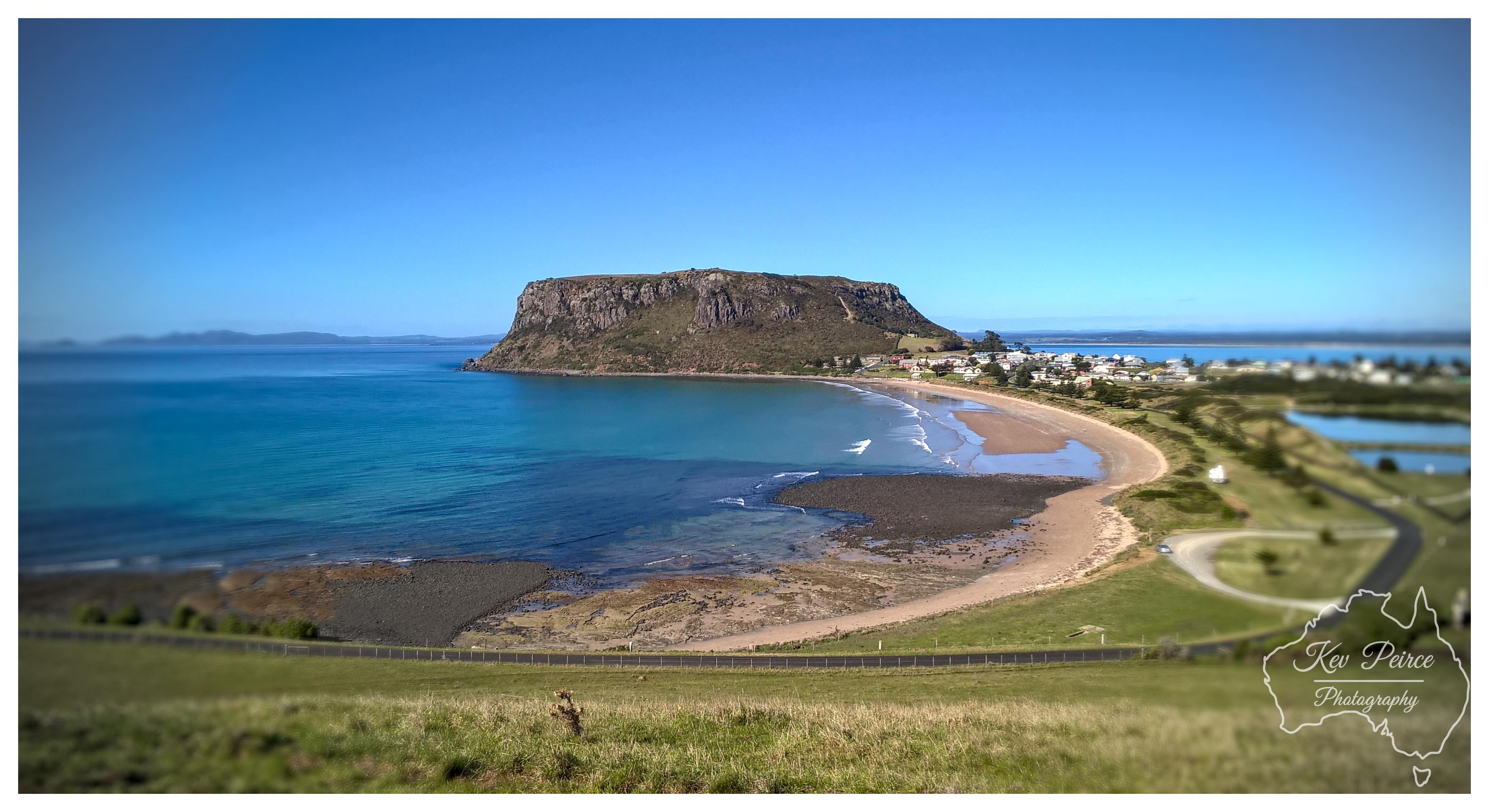 A wide elevated landscape shot capturing The Nut at Stanley rising dramatically above a sweeping curved bay and sandy shoreline. Bright midday sunlight turns the shallow coastal waters vibrant shades of turquoise and deep blue. A grassy hill and roadside fence sit in the foreground, leading down to the coastal township nestled against the base of the massive volcanic bluff.